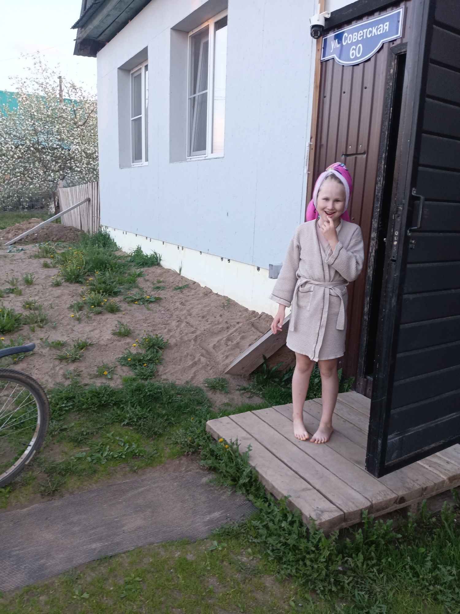 A young girl in a light brown robe and pink towel-wrapped hair stands on a wooden porch in front of a brown door. A street sign reading "ул. Советская 60" is above the door, identifying the location as 60 Sovetskaya Street. The area in front of the house is sandy with patches of grass and dandelions, and a bicycle wheel is visible on the left. The building is light blue, with two windows, and a tree with white blossoms is in the background. The city is identified as Фоки, Russia.