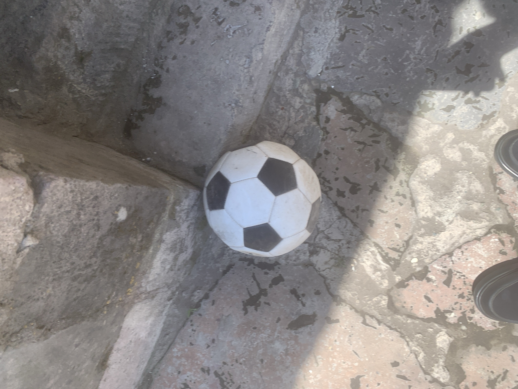 The image captures an outdoor scene in Gyumri, Armenia, viewed from a high angle. A worn, black and white soccer ball, appearing somewhat misshapen or partially deflated, rests in the corner formed by two converging rough, grey stone or concrete structures, likely steps or low walls. The ground beneath the ball and extending into the foreground consists of irregular, weathered stone or concrete pavers, displaying a mix of reddish-brown and grey tones, along with visible cracks and dark, irregular stains. Strong sunlight illuminates the scene, casting distinct, elongated shadows across the ground and portions of the stone structures. In the bottom left and right quadrants of the frame, the dark-colored shoes of an individual are partially visible, indicating a human presence observing the stationary ball and environment. No other individuals or active interactions are present.