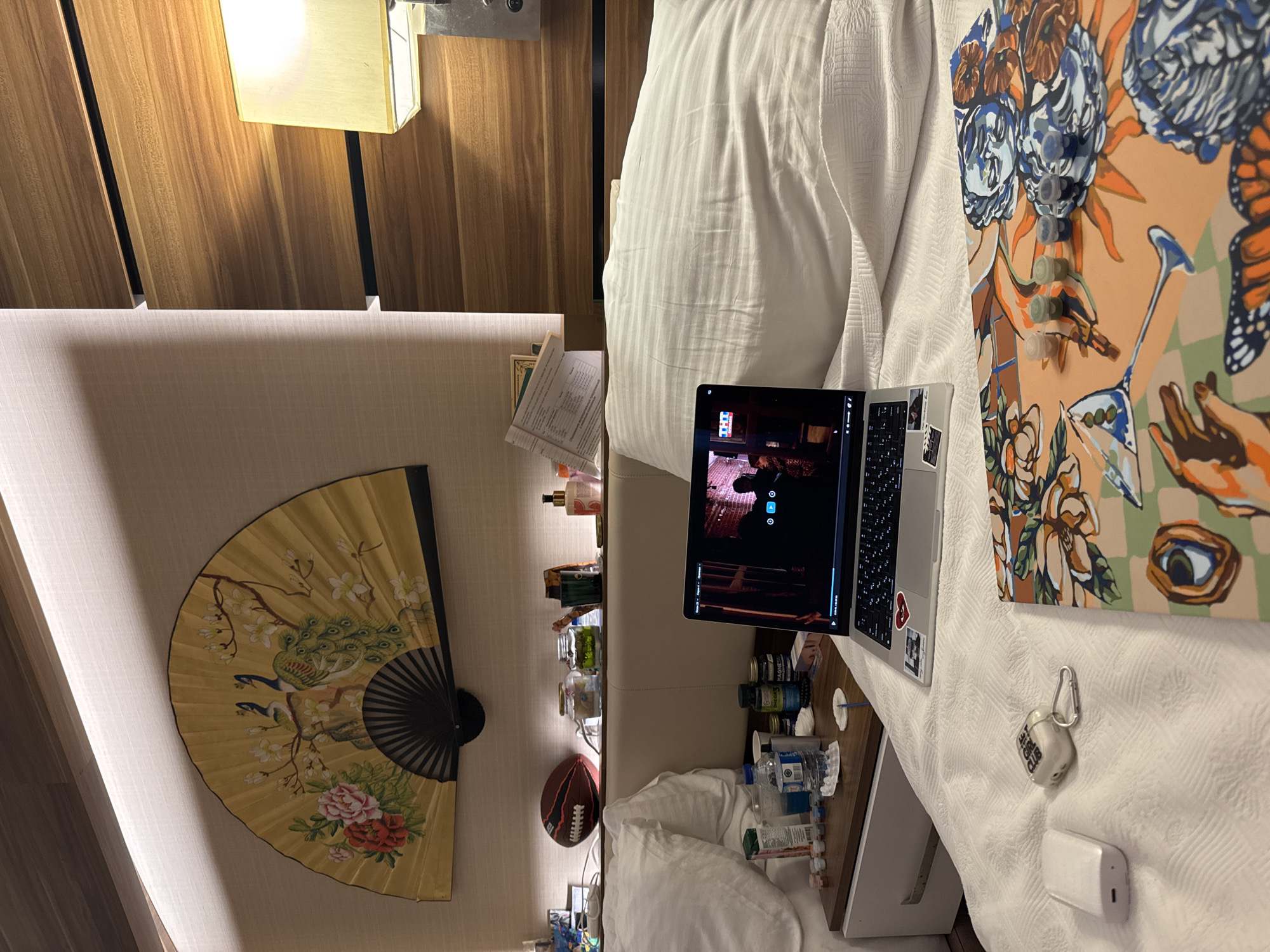 The image depicts a bedroom scene, likely in a hotel, featuring a neatly made bed with white striped bedding. On the bed, a laptop is open and displaying a video with a thumbnail that includes the "THC" logo. To the left of the laptop, on a nightstand, are various items including medicine bottles, a water bottle, and a small white box. Further to the left on the nightstand, a football is visible. Above the nightstand, a decorative fan with a peacock and floral motif is mounted on the wall. To the right of the bed, a wooden headboard is visible, with a lampshade casting a warm light. In the foreground on the bed, there is an open paint-by-numbers canvas with small pots of paint. Also on the bed are a white wireless earbud case with a carabiner clip and a small white rectangular electronic device. The overall impression is one of a relaxed evening setting. The location in the city of Aksu, Turkey, is not discernible from the visual elements present in the image.