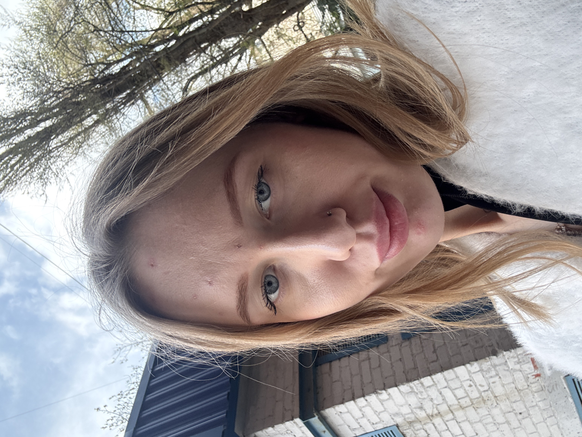 A low-angle selfie of a young woman with blonde hair, fair skin, and blue eyes. She has a nose piercing and some blemishes on her forehead. A tree with bare branches is visible in the upper left corner, with a cloudy sky in the background. The lower portion of the image shows a brick building and a corrugated metal structure. The location appears to be outdoors. There's no information about Rostov-on-Don.