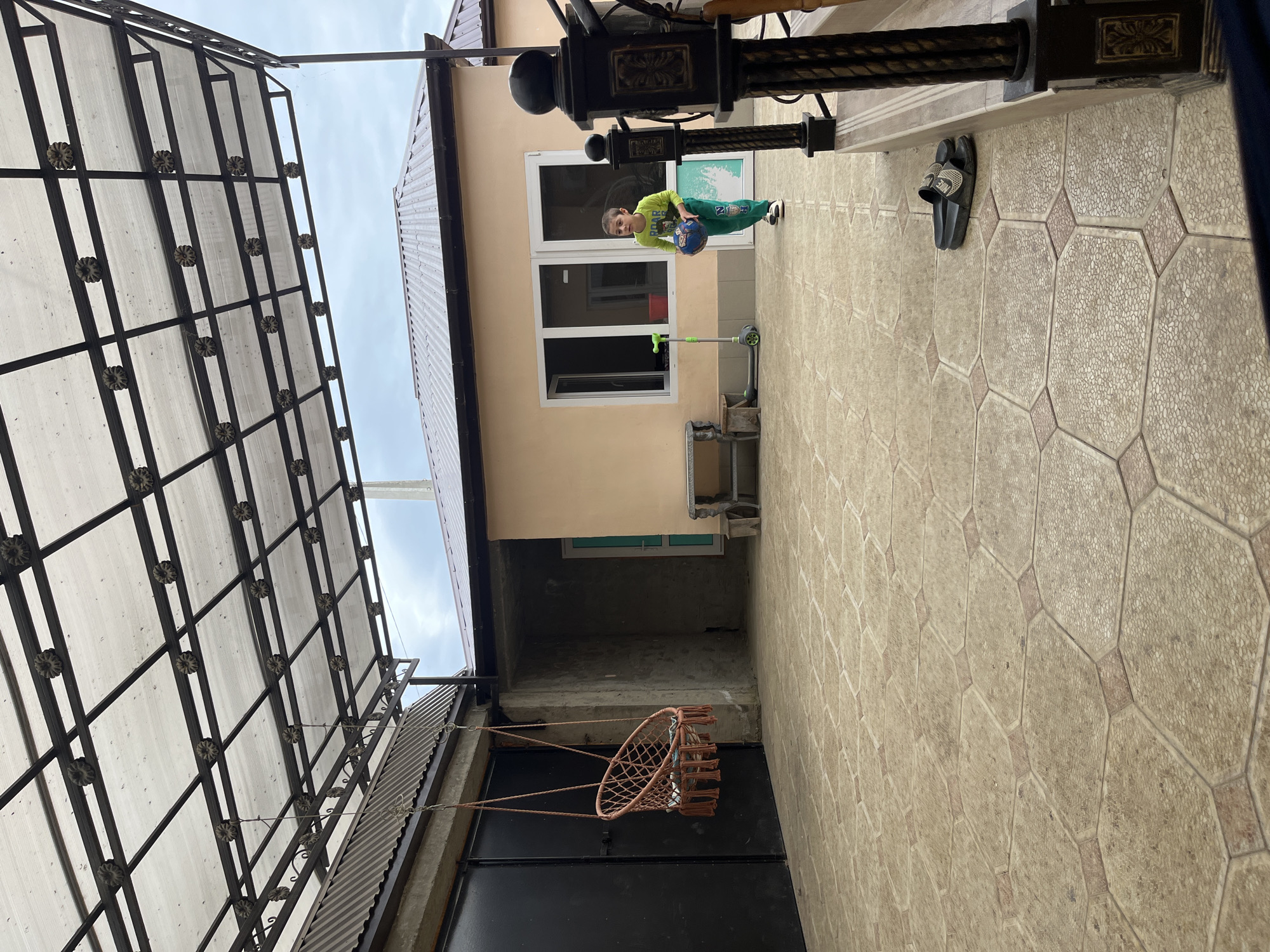 A full shot captures a young boy in a green shirt and pants, holding a blue soccer ball, standing in a courtyard. A dark-colored scooter is visible near him. Behind the boy is a two-story building with a beige facade and white-framed windows. A basketball hoop hangs from a garage door under a metal-framed canopy on the left. The courtyard floor is tiled with hexagonal patterned paving stones. Location: Kaspiysk, Russia.