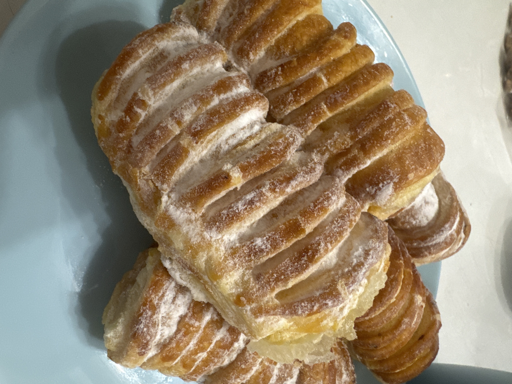 A close-up, overhead shot shows a light blue plate filled with several golden-brown pastries dusted with powdered sugar. The pastries have a distinct ridged pattern on top and appear to be a sweet bread or Danish. The background is blurred, suggesting an indoor setting, possibly a cafe or home kitchen. There is no indication of the location being Sochi, Russia, nor are there any people or actions depicted.