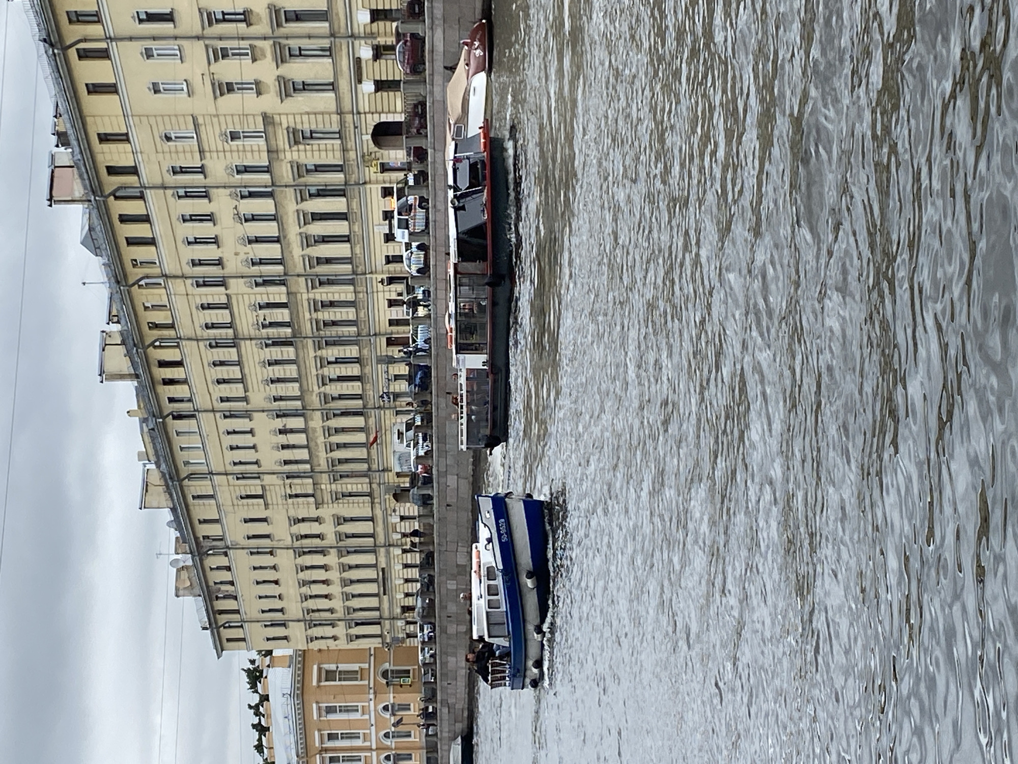 The image shows a canal in Saint Petersburg, Russia, with a beige, multi-story building lining the embankment. The building has numerous windows and architectural details typical of 19th-century St. Petersburg. In the foreground, two boats are visible on the canal. The boat on the left is smaller, with a blue and white hull, and appears to be a passenger boat with several people on board. It has the number "50-0629" on its side. The boat on the right is larger and longer, with a darker hull and a cabin. It also seems to be carrying passengers. In the background, a large building complex stretches along the embankment, with trees and more buildings visible behind it. The sky is overcast, suggesting a cloudy day. The water is choppy, indicating movement.