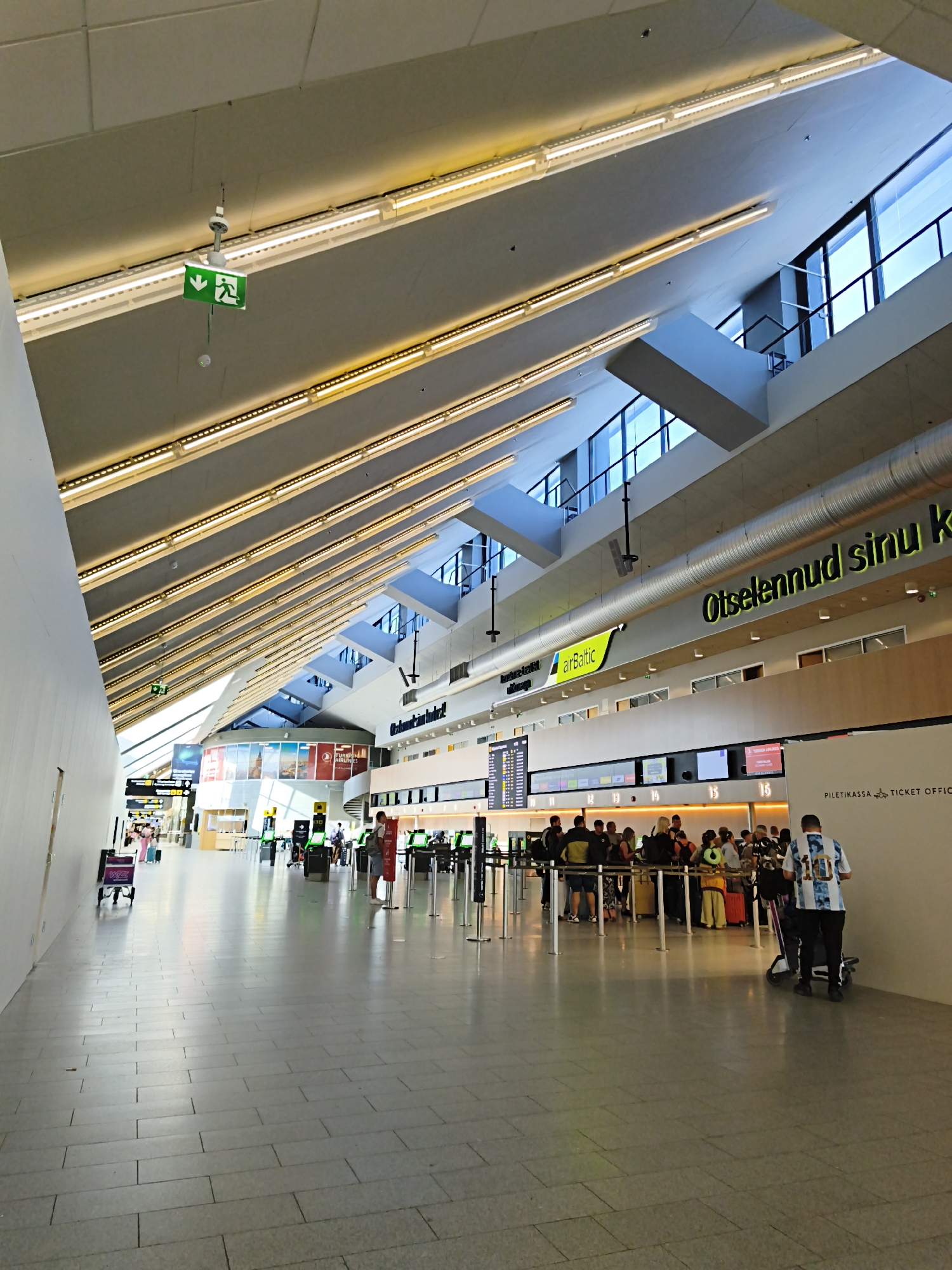 The image depicts the interior of Tallinn Airport (TLL) in Tallinn, Estonia. The scene is an airport terminal with a high, sloped ceiling featuring numerous light fixtures and a large skylight allowing natural light to enter. In the foreground, a wide tiled walkway is visible. Further into the terminal, a queue of travelers is lined up at check-in counters. Above the counters, a large digital display board shows flight information. Several self-service check-in kiosks are also present. A prominent sign with green text on a yellow background reads "airBaltic." Another sign in large yellow letters on the ceiling states "Otselennud sinu k." People are seen with luggage, some pulling suitcases and others with carry-on bags. An exit sign with a running man icon is suspended from the ceiling. The overall atmosphere suggests a busy travel hub.