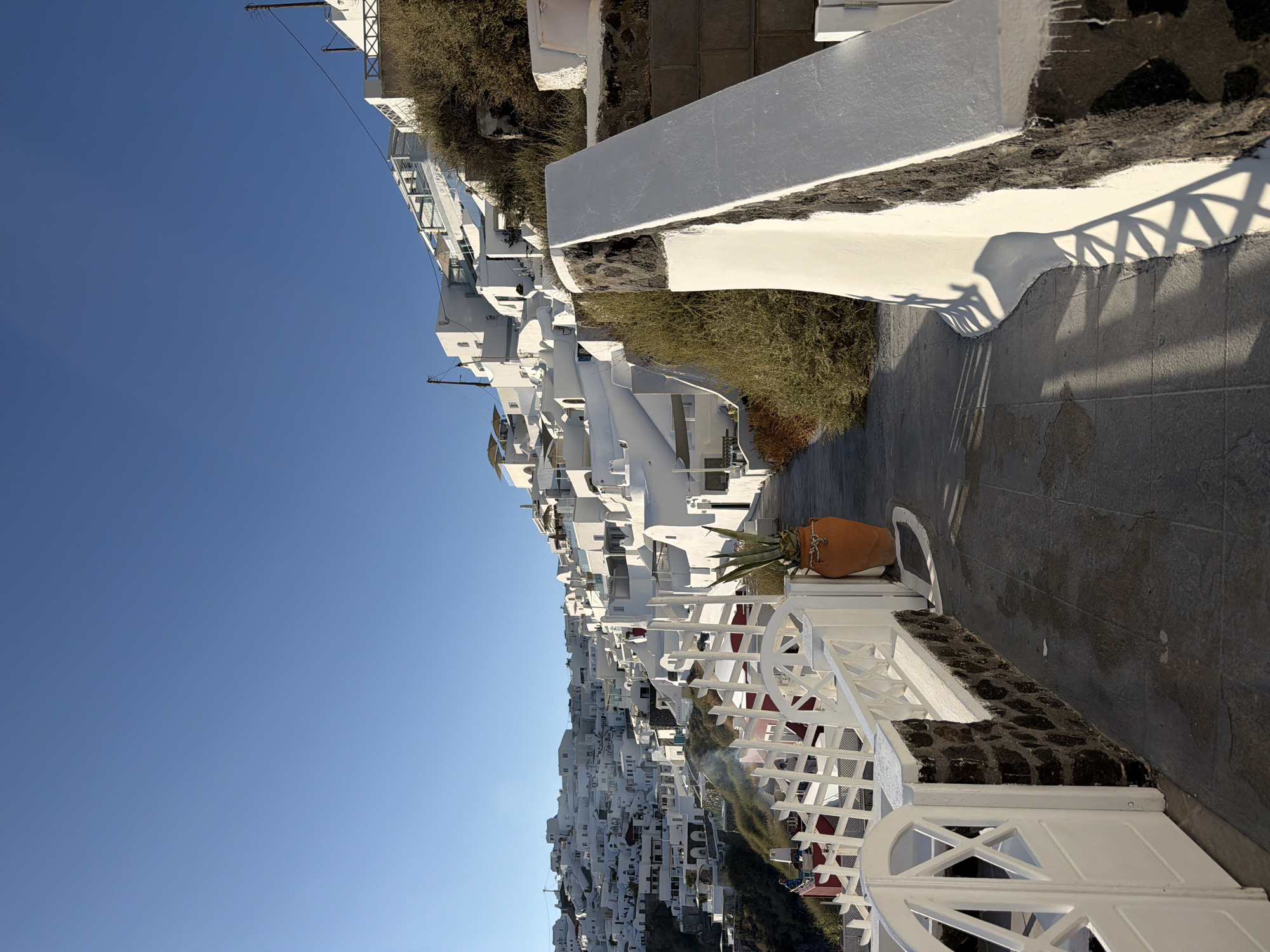 The image depicts a scenic view of a hillside town with whitewashed buildings against a clear blue sky. The architecture is characteristic of the Cycladic style, with cubical houses cascading down the slope. A paved pathway leads through the town, bordered by a white railing and dark volcanic stone walls. Lush green foliage grows on the hillsides, contrasting with the white structures. A large terracotta pot with a plant sits next to the pathway. In the background, more buildings are visible, some with balconies and terraces. The scene evokes a sense of tranquility and traditional Mediterranean charm, likely captured in the city of Thira on the island of Santorini, Greece, known for its dramatic cliffside villages and stunning Aegean Sea views. There are no people clearly visible in the frame.