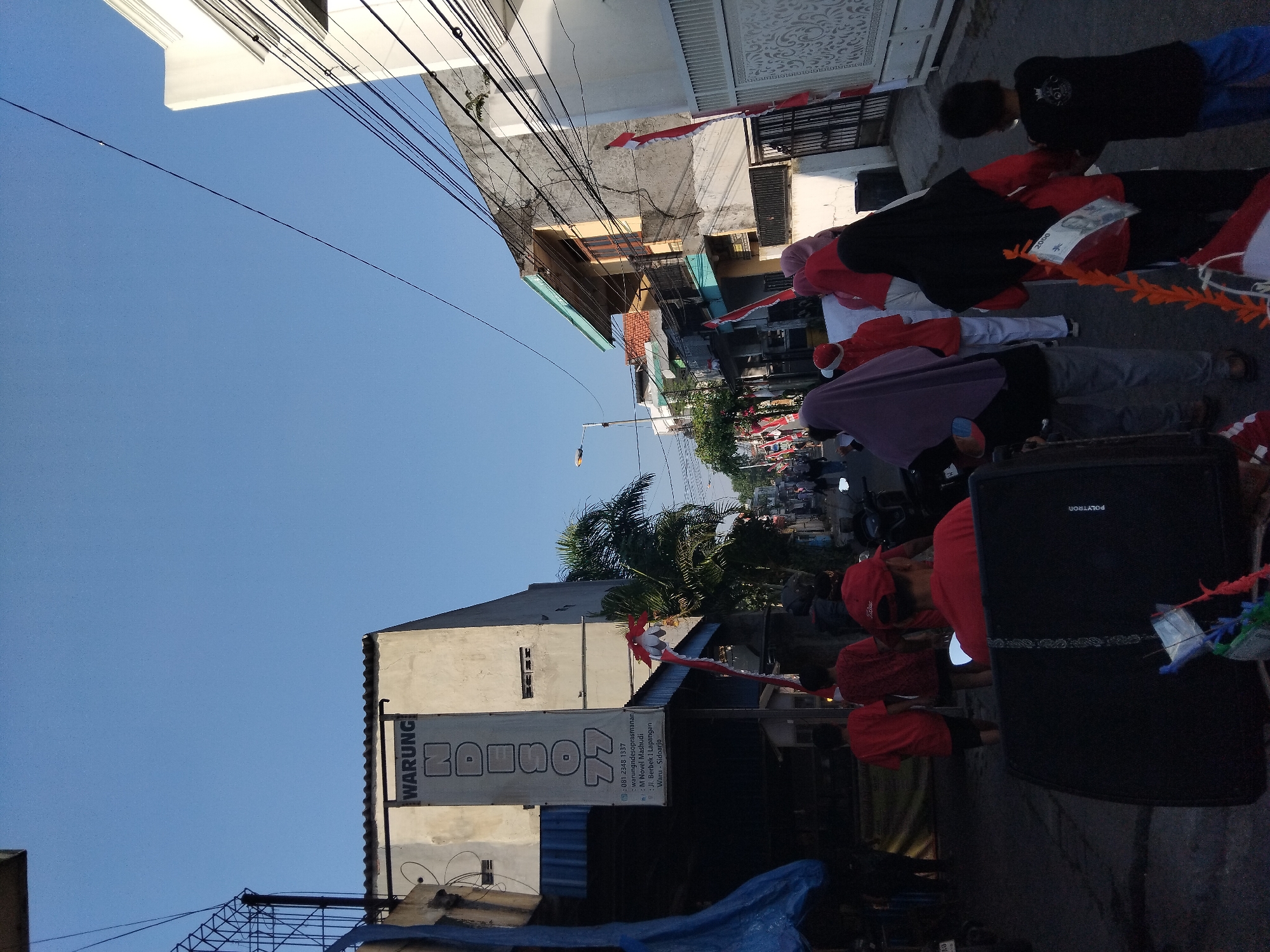 The image depicts a street scene in Berbek, Indonesia, likely during a public gathering or celebration, given the presence of Indonesian flags. In the foreground, a large black speaker is partially visible, with a person wearing a red shirt and cap facing away from the camera, seated behind it. To the left, several individuals in red shirts are walking away from the camera. In the mid-ground, a group of people, some wearing headscarves, are walking down the street. One person in a red shirt has a small Indonesian flag attached to their back. On the left side of the street, a building with a banner that reads "WARUNG NDESO 77" is visible, along with a telephone number and address. The street is lined with buildings on both sides, with power lines crisscrossing overhead. The sky is clear and blue, indicating a sunny day. The overall atmosphere appears to be one of community activity.