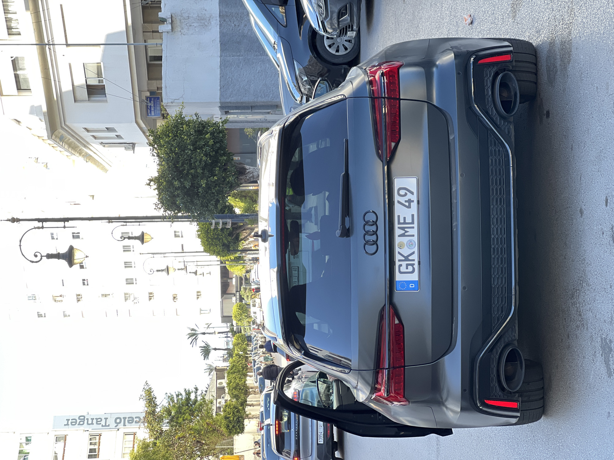 The image displays the rear view of a dark grey, satin-finish Audi RS6 Avant stationed on an asphalt road in Tanger, Morocco. The vehicle exhibits a German license plate, "GK ME 49", which includes the German flag and a circular emblem. The driver's side rear passenger door of the Audi is open.
To the left of the Audi, a silver Peugeot 3008 is visible with its rear hatch open, followed by a queue of other vehicles in what appears to be active traffic. Distant pedestrians are discernible on sidewalks. The street is bordered by multi-story white buildings. A vertical sign on a building on the left reads "Barceló Tanger", confirming the city. Ornate black street lamps are positioned along the thoroughfare. Green trees, including palm trees, and various bushes line the street. To the right, a blue sign on a building identifies "مستشفى مولاي الحسن" (Moulay El Hassan Hospital) and "Tanger". The scene is illuminated by clear daylight, indicating fair weather.