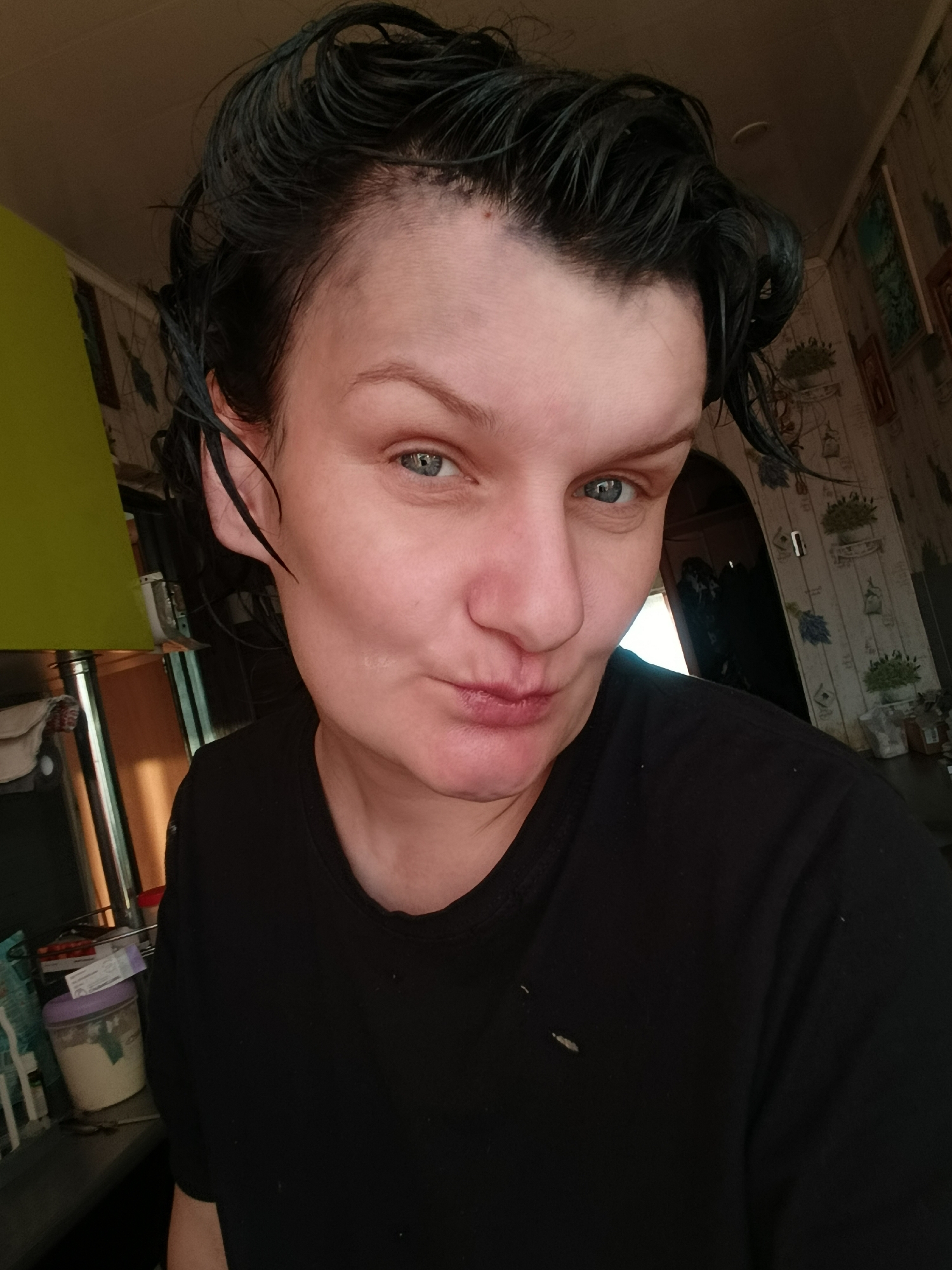 A selfie of a middle-aged woman with wet, dark hair, light skin, and blue eyes. She is wearing a black t-shirt. The background shows a room with patterned wallpaper, likely a kitchen, with some objects visible on a counter. Based on the prompt referencing Фоки, Russia, it's implied the location is within that city, though there's no explicit visual evidence to confirm this.