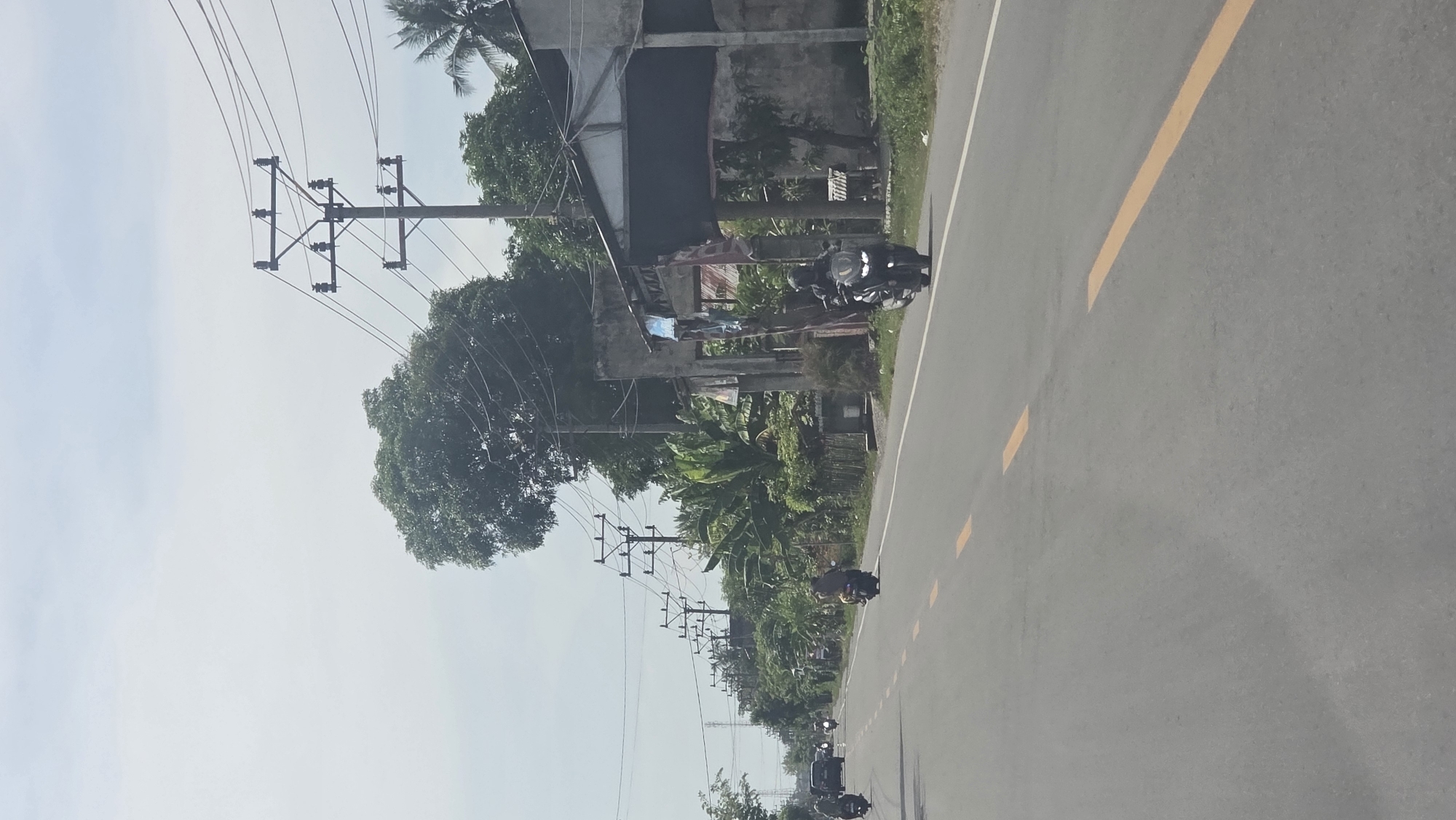 The scene depicts a road in Teungoh Baro, Indonesia. The road has a gray surface with yellow lane markings and is flanked by utility poles with overhead power lines. Several motorcycles are visible on the road, moving in the same direction. On the side of the road, there are trees, vegetation, and a building. Clothing is hanging on a line near the building. The sky is visible in the upper portion of the frame.