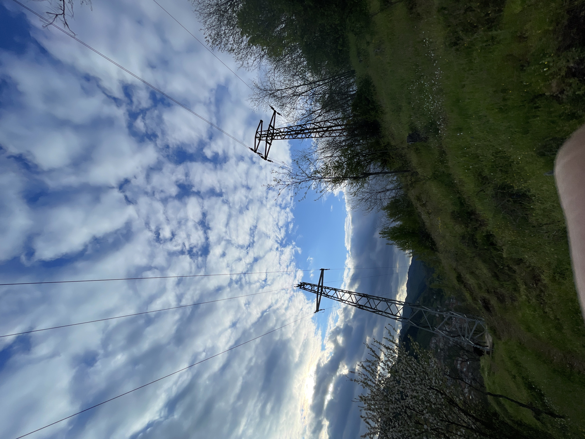 The image is taken from a low angle, looking upward toward two electrical transmission towers. The towers are constructed of a lattice of steel and support several high-voltage power lines. The sky is filled with clouds, with patches of blue sky visible between them. On the right side of the frame is a grassy slope, with trees and some flowering plants at the bottom. In the background, there appears to be a distant mountain range and a glimpse of a settlement. The image likely captures a landscape near Zlatograd, Bulgaria, where similar terrains and transmission infrastructure are located.