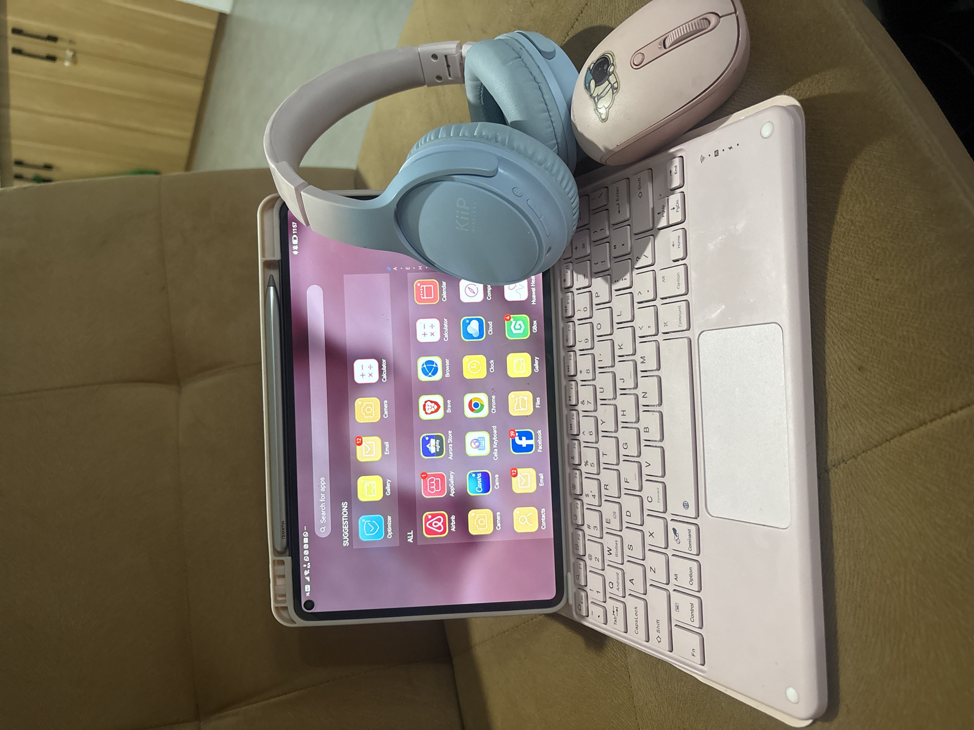 An overhead, slightly angled shot captures an electronic device setup on a textured, dark brown or tan upholstered surface, likely a couch. The primary subject is a tablet integrated into a light pink keyboard case, positioned horizontally. A silver stylus is docked on the left side of the tablet. On the screen, an app drawer is displayed with numerous app icons, a "Search for apps" bar, and "SUGGESTIONS." Visible app names include "Optimizer," "Gallery," "Email," "Camera," "Calculator," "Airbnb," "AppGallery," "Aurora Store," "Brave," "Browser," "Calendar," "Canva," "Celia Keyboard," "Chrome," "Clock," "Cloud," "Files," "Facebook," "GBox," and "Huawei Health." Notifications like "12" for Email and "39" for Facebook are present, and the time "11:57" is displayed.
Resting on the tablet's upper right corner and extending onto the keyboard is a pair of light blue over-ear headphones, bearing the text "KiiP WIRELESS" on the earcup. To the right of the headphones, partially on the keyboard, is a light pink wireless mouse adorned with a small astronaut sticker.
The keyboard, matching the light pink of the case and mouse, features a standard QWERTY layout. Notable keys include "Android," "Windows," "IOS," and "Connect." A touchpad is integrated below the spacebar. The setting appears to be indoors with ambient lighting.