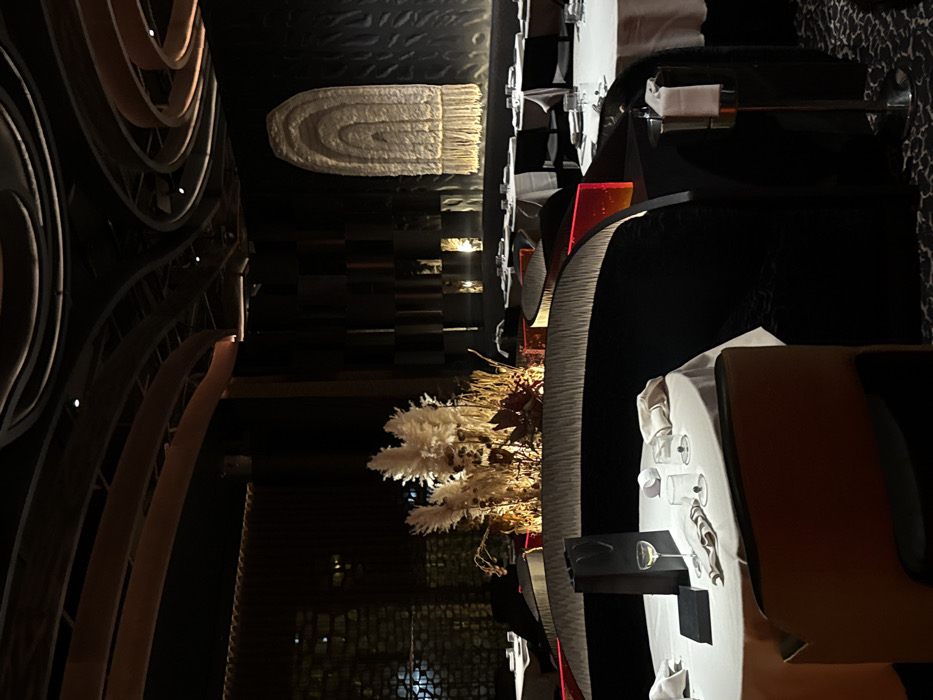 A dimly lit, stylish restaurant interior features black chairs and tables elegantly set with white linens and glassware. A central floral arrangement sits amidst the tables. Overhead, a modern art installation complements a textured wall. The ceiling is decorated with an unusual, woven pendant lamp. The scene exudes an ambiance of sophistication and understated luxury.