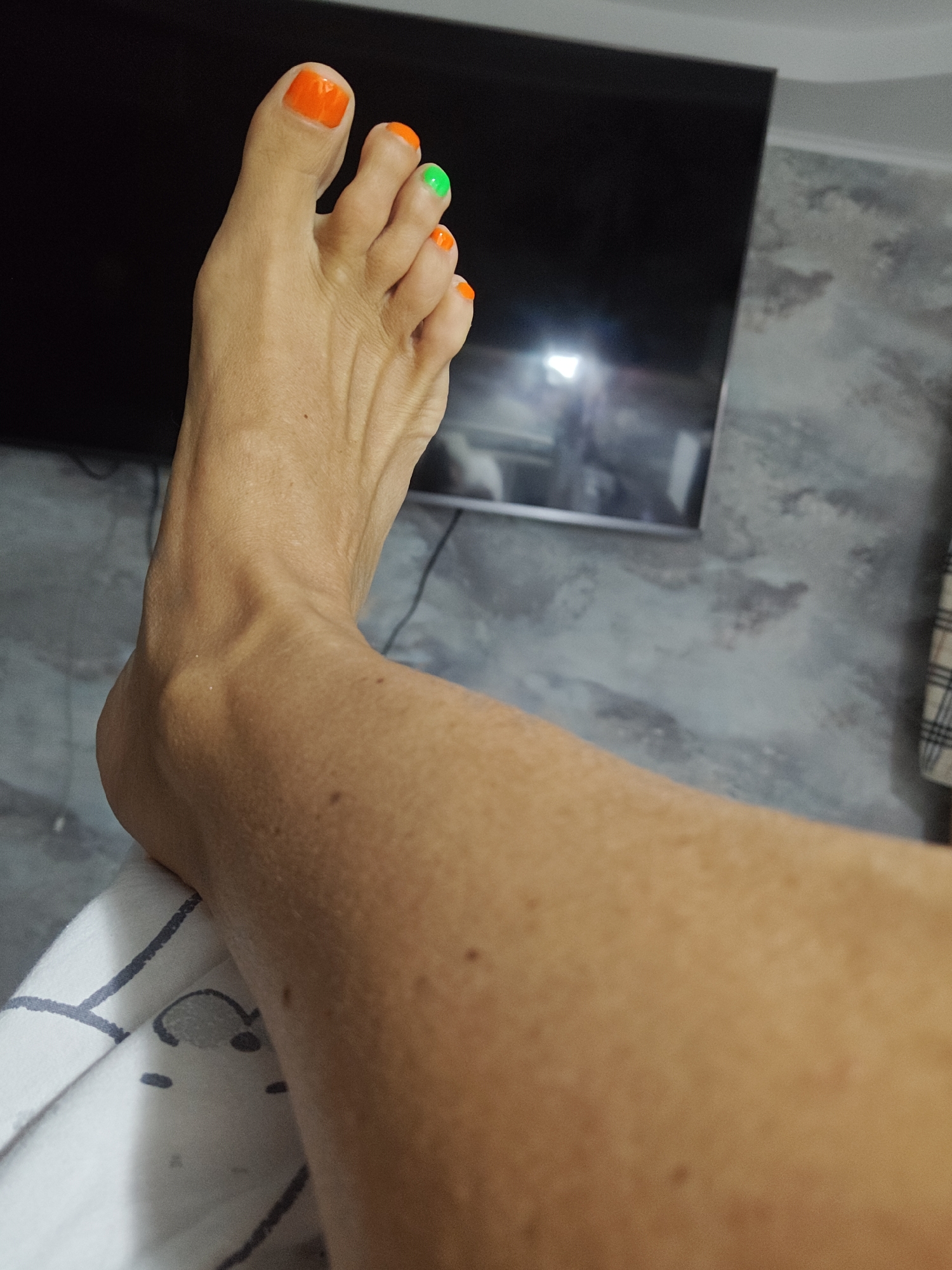 A close-up, oblique, eye-level shot focuses on a person's left leg and foot, which are positioned in the foreground. The leg is extended and draped over a white fabric with a dark grey striped pattern. The skin of the leg is tanned and has visible pores and moles. The foot is raised, with toes adorned with orange nail polish on the big toe and smallest toe, and green nail polish on the second toe. The background features a black television screen and a grey, textured wall. The scene appears to be indoors. The specific location in Komsomolsk-on-Amur, Russia, is not discernible from the visual information provided in the image.
