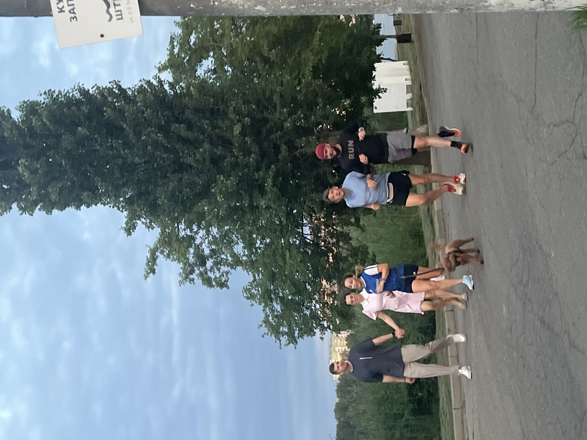 In Krasnodar, Russia, a group of five people and a small brown poodle are jogging on a paved path. The sky is overcast with light clouds. On the right side of the image, a utility pole with a white sign is visible. The sign has Cyrillic text and a drawing of a snake. The people are dressed in athletic wear, except for one woman in a pink dress. They are running in a line, with the man in the pink dress and the woman in the blue shirt closest to the camera. The poodle is running ahead of the group. The background consists of trees and some buildings in the distance.
