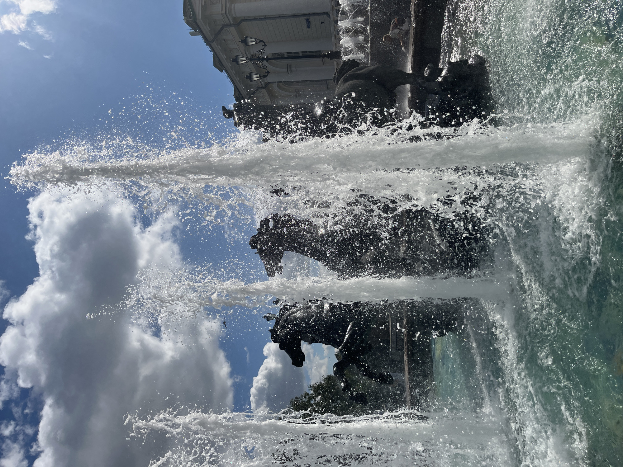 This image depicts the Fountain of the Four Seasons, located in the Alexander Garden in Moscow, Russia. The fountain features a central sculpture of four horses, each representing a season, with water jets spraying upwards. In the foreground, several large streams of water from the fountain are prominent, obscuring parts of the sculpture and the background. The water in the fountain basin appears clear and turquoise. Behind the fountain, a building with classical architecture is visible, and to the right, a lamppost with three lights stands. A few people are present near the fountain, one of whom appears to be sitting by the edge. The sky is a bright blue with scattered white clouds, and the sun is casting light and shadows, creating a sparkling effect on the water. Lush green trees are also visible in the background.