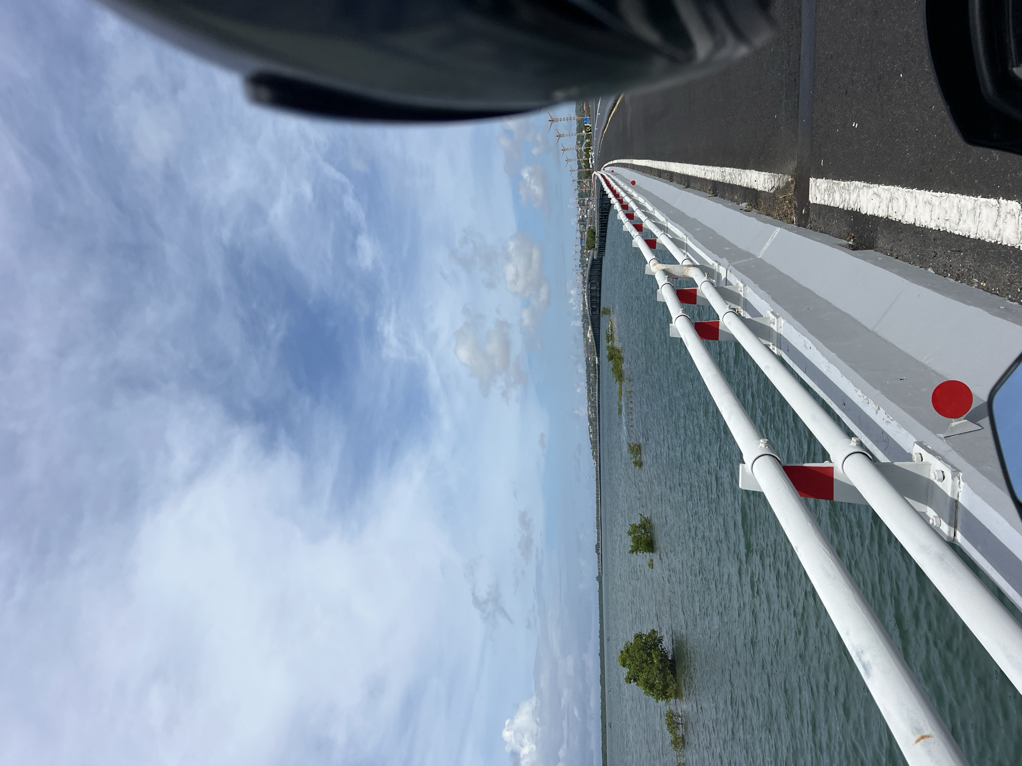 The image shows a view from a vehicle driving on a bridge. The bridge has white and red railings on one side, with a body of water visible below. In the distance, there appears to be a cityscape with construction cranes. The sky is blue with scattered clouds. The vehicle's interior is partially visible in the top and right edges of the frame. Given the context and features, the location is likely on a bridge in Kuta, Indonesia, considering the coastal environment, presence of mangrove-like vegetation in the water, and construction activity in the background.