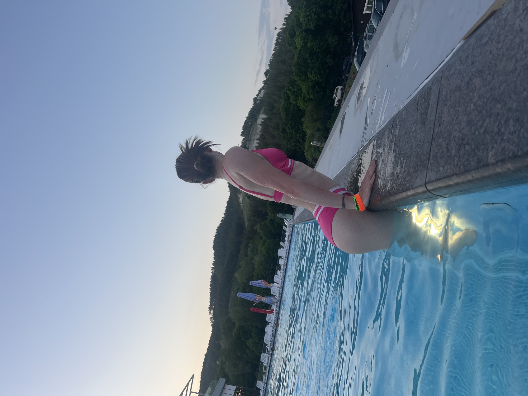 A woman in a pink bikini is at the edge of a swimming pool in what appears to be the city of Khvalynsk, Russia. She is holding onto the pool's edge. In the background is a wooded hillside and a row of parked cars. Lounge chairs and umbrellas are visible next to the pool. The sky is a clear, pale blue.