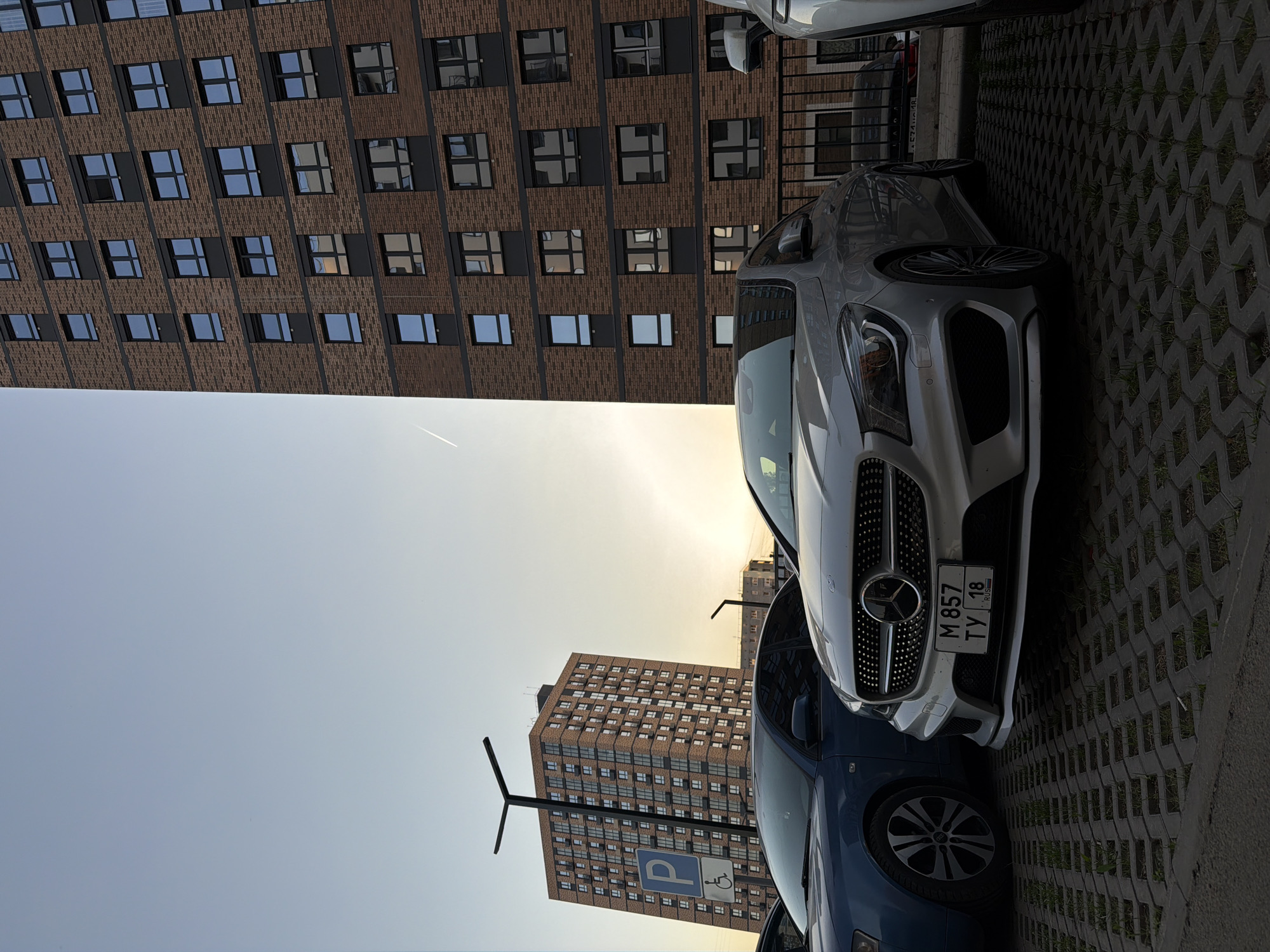 The image shows a street scene in Izhevsk, Russia. Two multi-story brick buildings dominate the background, one taller than the other. The sky is a pale yellow near the horizon, transitioning to a light blue higher up. In the foreground are two cars parked on a grey interlocking brick surface. The silver car in the center has a Russian license plate that reads "M 857 TY 18 RUS." A blue car is partially visible beneath the silver car. A parking sign, illuminated by the sun, is visible near the bottom of the frame, indicating handicapped parking.