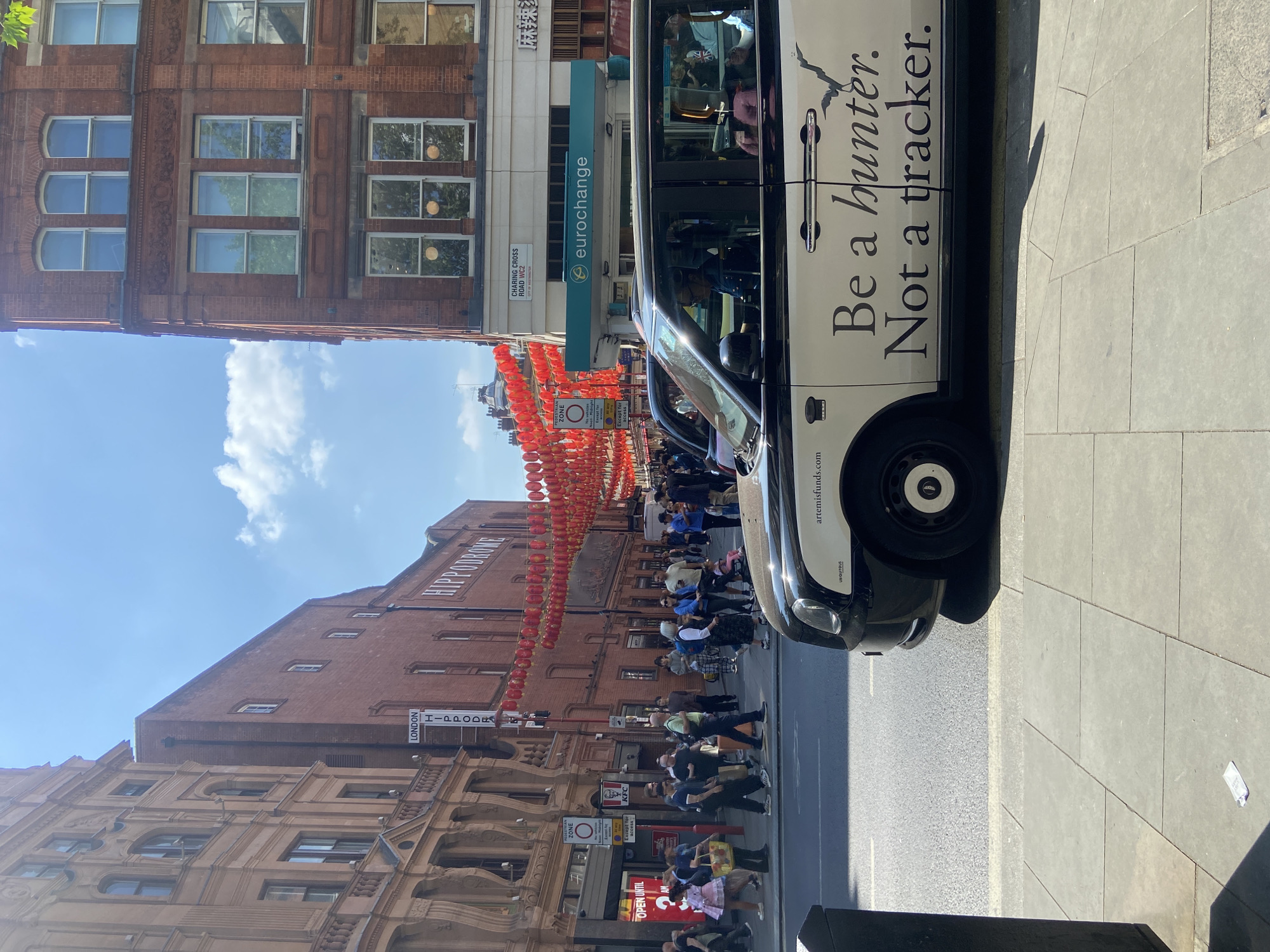 Here is a description of the image:
This image captures a street scene in London's Chinatown, in the United Kingdom. In the foreground, a black and white taxi cab parked on the side of the street obscures part of the view. The taxi has the words "Be a hunter. Not a tracker." written on it.
The backdrop is dominated by traditional brick buildings, some bearing ornate architectural details. A prominent building is the London Hippodrome, with signage visible. Strings of vibrant red Chinese lanterns stretch across the street, adding to the area's distinctive aesthetic.
Several pedestrians can be seen walking along the pavement and crossing the street. Signage for various businesses, including Eurochange and KFC, is visible on the buildings. The sky is partly cloudy, suggesting a pleasant day. Street signs indicate that the location is Charing Cross Road.