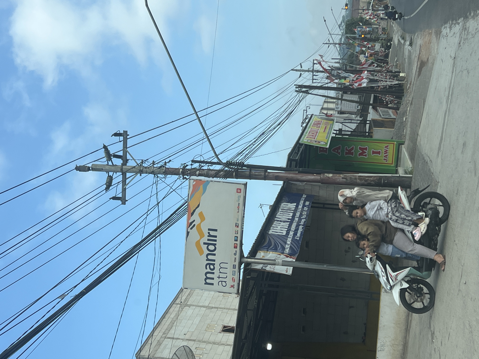 The image captures a street scene in Kranggan, Indonesia. In the foreground, a white scooter carries four people. A woman is driving, with three children seated behind her. The children appear to be of varying ages, with one in front of her and two behind. The scooter is positioned on a paved road.
To the left of the scooter is a building with a white facade and a sign that reads "mandiri atm". Below this sign, a smaller banner displays "DHR MOTOR". Adjacent to this is a green building with a sign reading "BAKMI JAWA".
The environment is characterized by numerous overhead power lines, some of which are supported by wooden utility poles. Along the side of the road, there are many Indonesian flags displayed on poles, suggesting a festive occasion or a public display. Further down the road, other buildings and more flags are visible. The sky is bright blue with some scattered clouds. A few motorcycles can be seen further down the road, suggesting ongoing traffic.