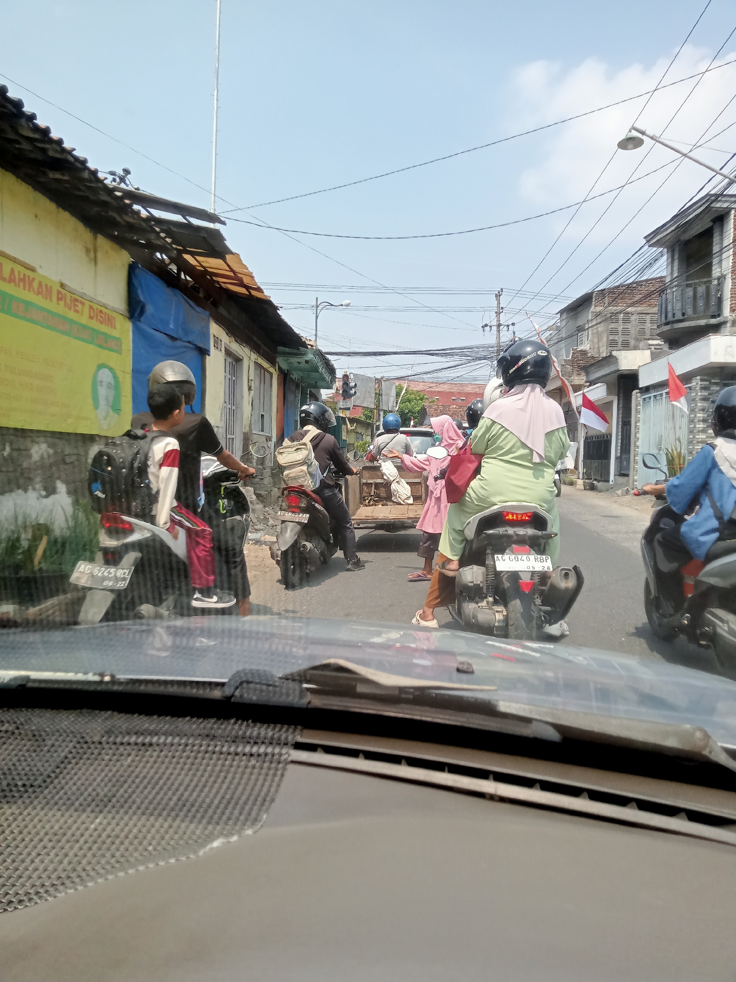 In Mangunsari, Indonesia, on a bright, sunny day, the scene captured from within a vehicle shows a street with several motorcycles and their riders. The foreground is dominated by the interior of a car, specifically the dashboard and windshield, which are slightly blurry. Beyond the windshield, a white motorcycle is parked on the left, with a young boy wearing a backpack and a striped shirt seated on it. To his right, a person wearing a black shirt and helmet is seated on the same motorcycle.
In the middle ground, a light green motorcycle carries two individuals. The rider wears a light pink hijab and a light green garment, while a passenger behind her wears a pink hijab and a pink outfit, carrying a red bag. Both are wearing helmets. Further down the street, another motorcycle with a rider wearing a blue shirt and a helmet is visible. The street is lined with buildings on both sides, characterized by various architectural styles, some with visible signs and awnings. Overhead, a complex network of electrical wires crisscrosses the sky, with a lamppost and a tall antenna structure also present. Red and white Indonesian flags are displayed on some buildings, suggesting a national holiday or celebration. The road surface appears to be asphalt, and the general environment suggests a bustling urban or semi-urban setting.
