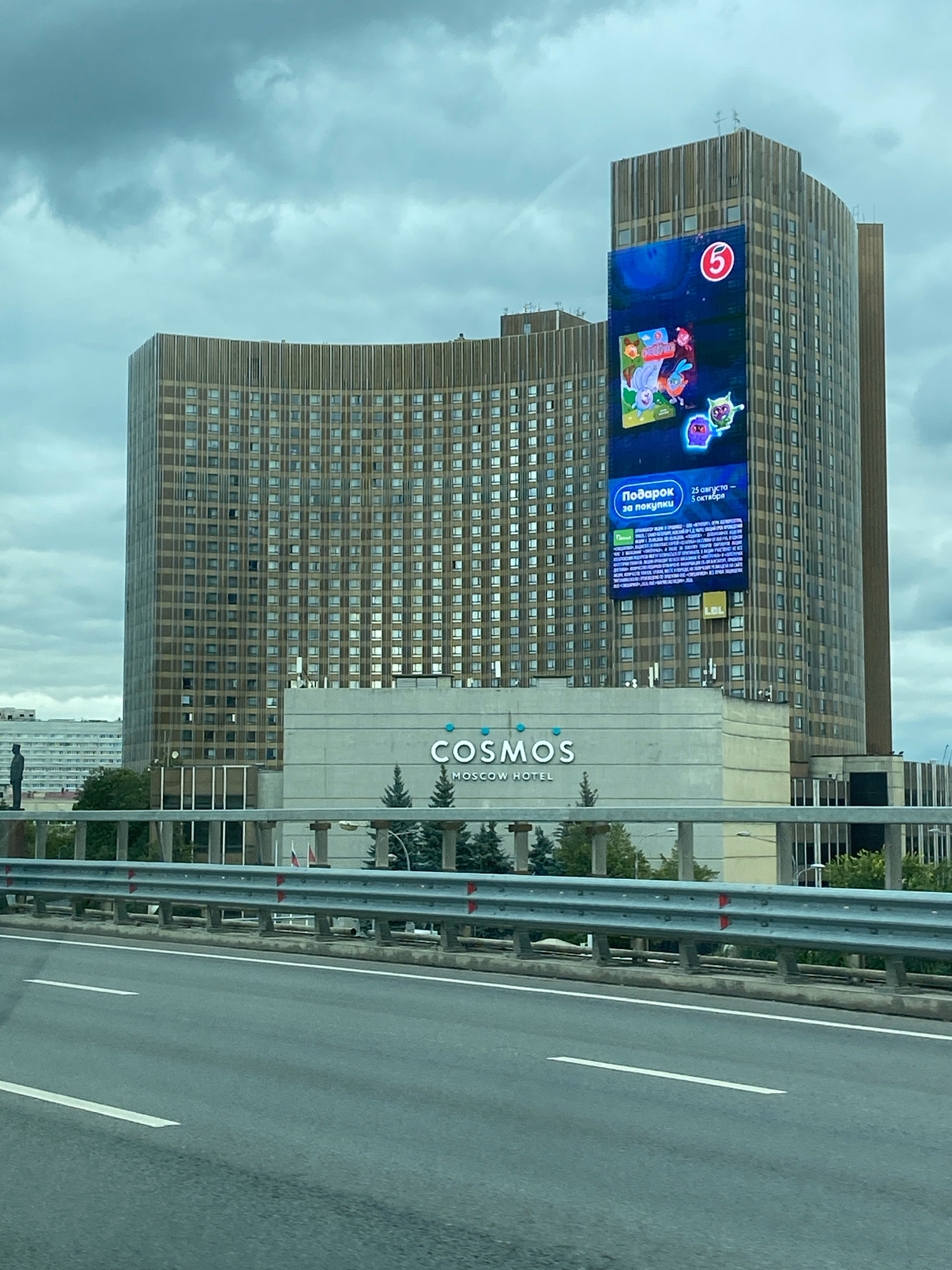 The image captures a large, curved hotel building in Moscow, Russia, identified by the "COSMOS MOSCOW HOTEL" signage on its lower section. A prominent digital billboard is mounted on the right side of the hotel, displaying vibrant cartoon characters and text in Russian, including "Подарок за покупки" (Gift for purchase) and a date range. The hotel is situated next to a highway, with a metal guardrail and traffic lanes visible in the foreground. The sky is overcast, with heavy clouds contributing to a muted light. A dark silhouette of a statue or monument can be seen on the left side of the frame, in front of a lower, lighter-colored building. Trees are planted in front of the hotel, and a few flagpoles are visible behind the guardrail. The overall scene depicts an urban environment with a focus on the architectural presence of the hotel and its advertising.