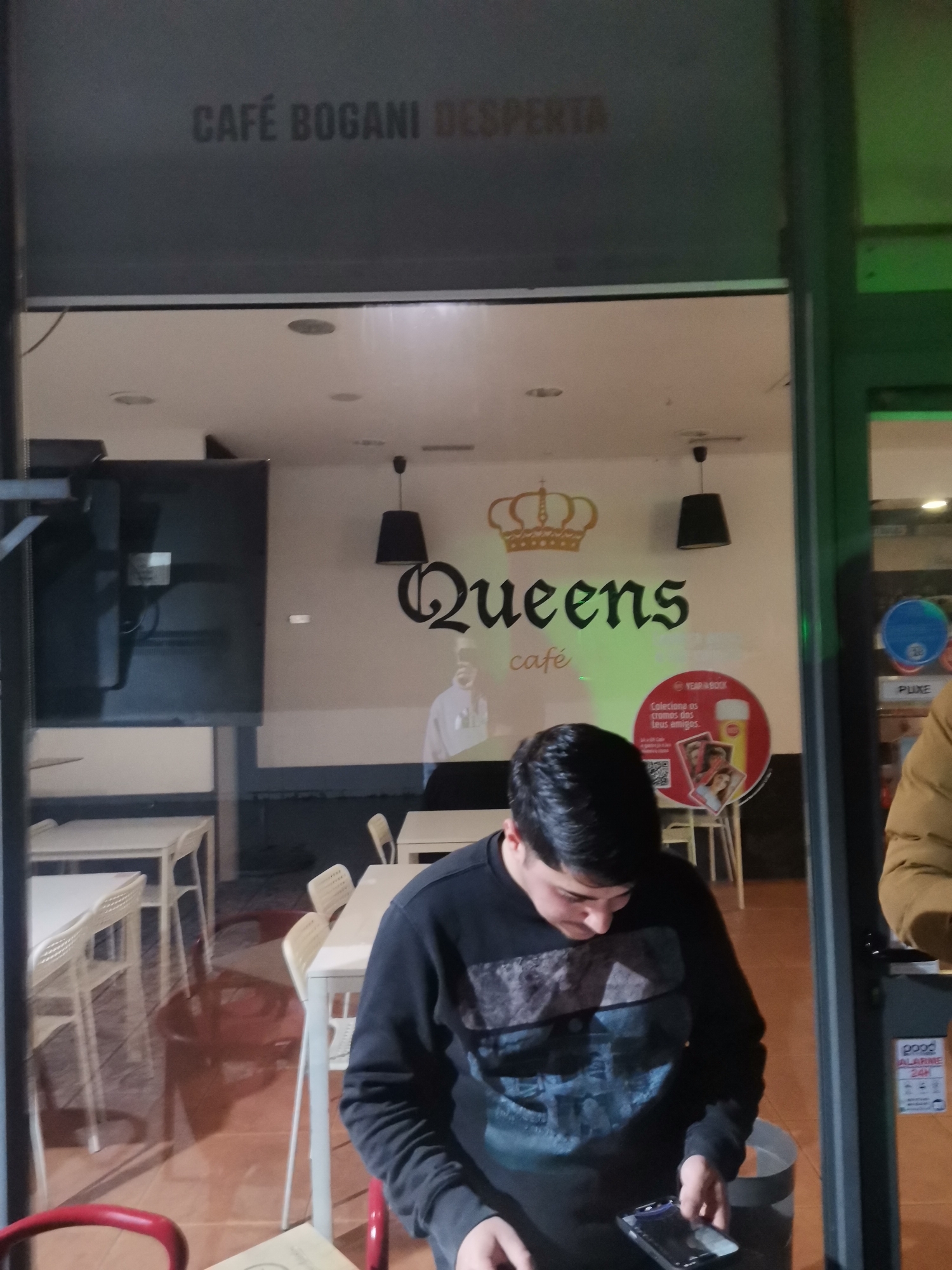 The image shows a person standing in front of a cafe named "Queens café," located in an unknown city. The cafe has a sign above the door that reads "CAFÉ BOGANI DESPERTA." Inside the cafe, there are tables and chairs, and a large screen television. There is a promotional poster about "cromos" on the wall. The person in the foreground is looking down at a phone in their hand. A part of another person is visible on the right side of the frame.
