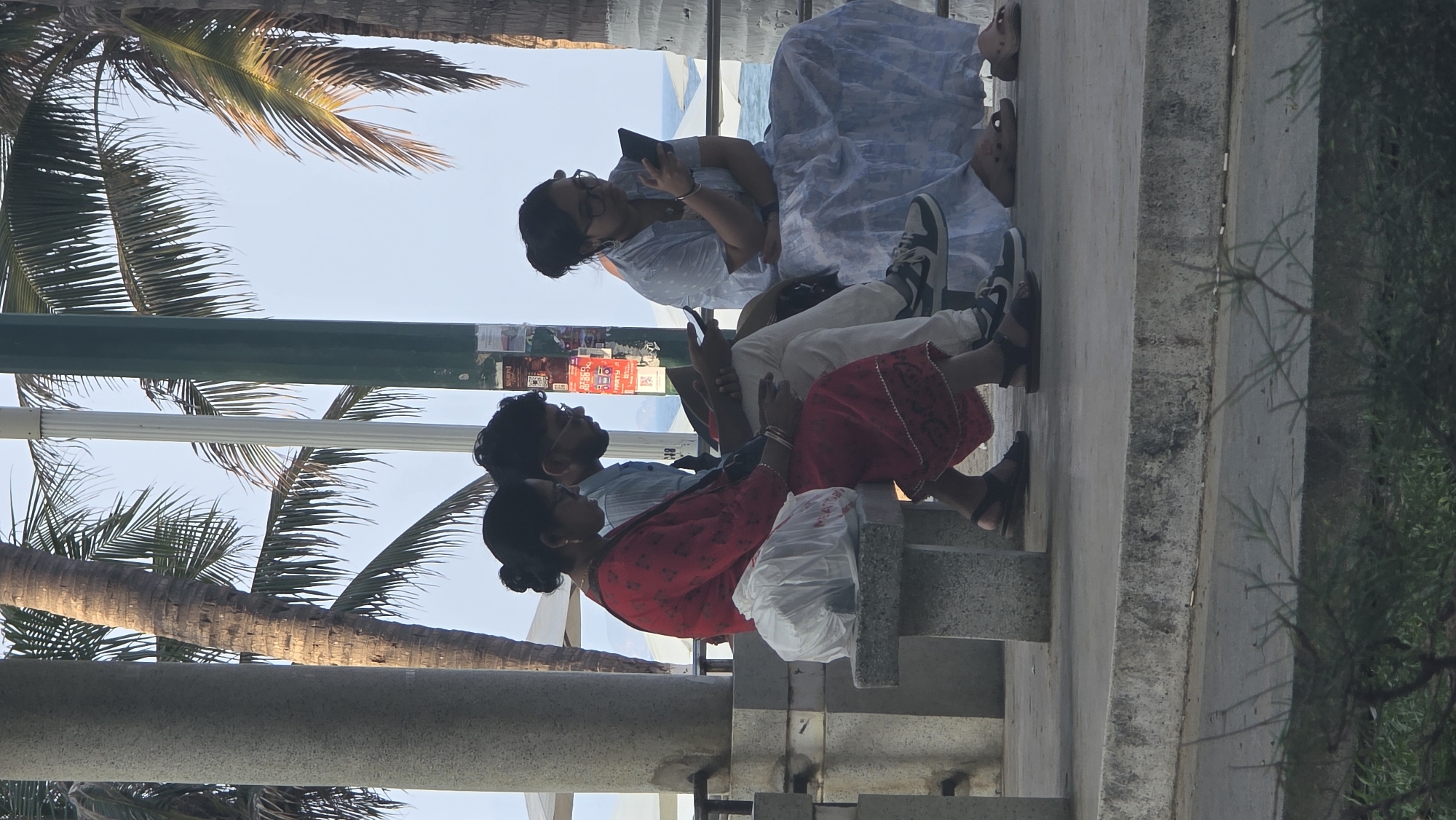The image captures a scene in Nha Trang, Vietnam, likely along a coastal area based on the presence of palm trees and possible view of the ocean in the background. Three individuals are seated on a concrete structure, possibly a bench or wall.
A woman in a light-colored, patterned dress is sitting towards the upper part of the image and looking at a phone in her hands. Another person, possibly a male, is sitting beside her, wearing light-colored pants. Closer to the bottom of the image, there is a man wearing a light blue shirt and beside him a woman in a red, patterned outfit. A plastic bag is placed on the concrete bench. They are all facing the same direction, possibly towards the water. The area is shaded by palm trees.