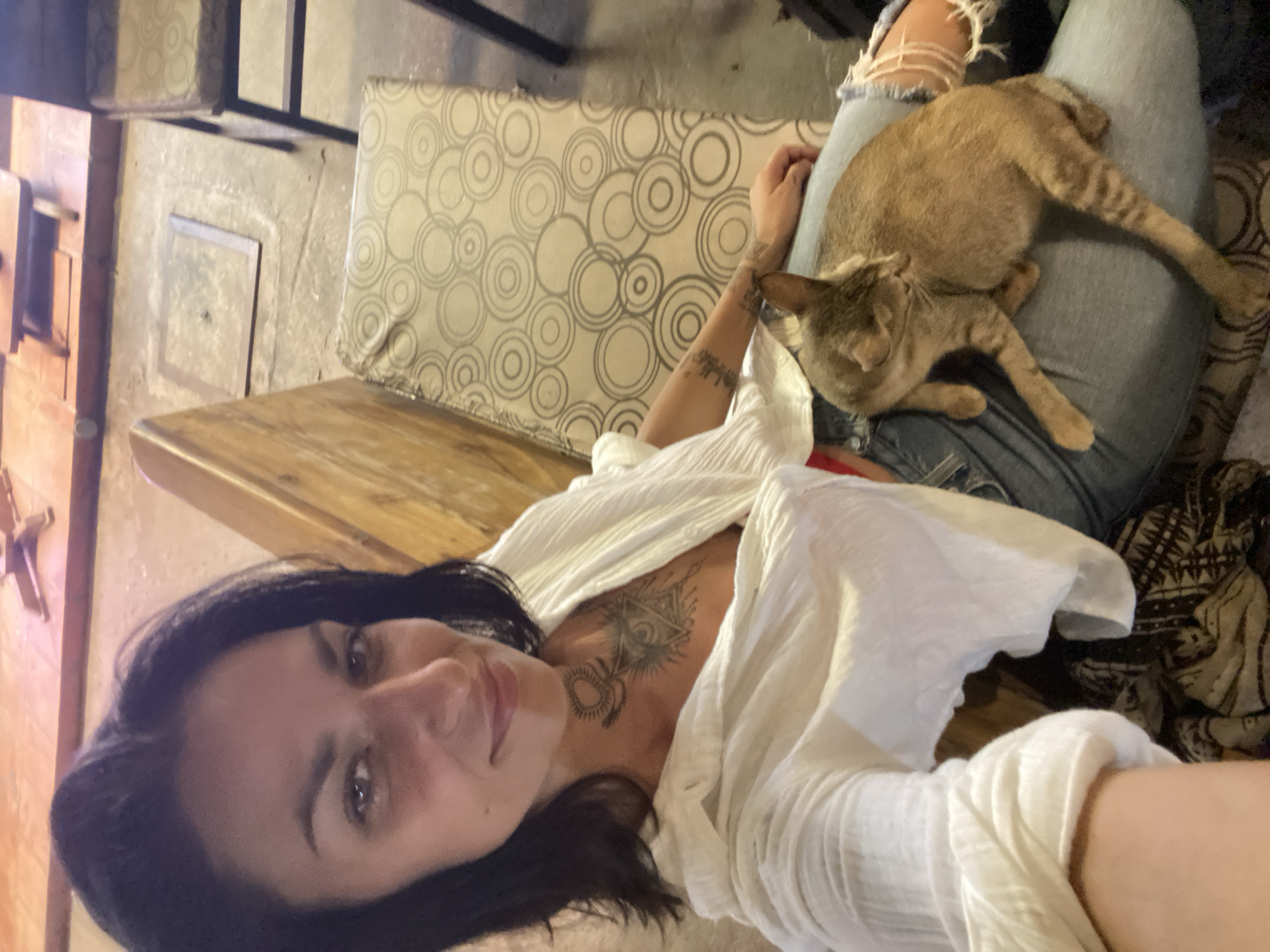 A young woman with dark hair and tattoos on her chest and arm is seated on a bench with a cat on her lap. She is wearing a white, open-collared shirt and ripped blue jeans. The cat is light brown with darker stripes. The background shows a concrete floor with a metal grate and wooden seating. The scene appears to be indoors, possibly in a restaurant or cafe, located in Phu Thuy Ward, Vietnam.