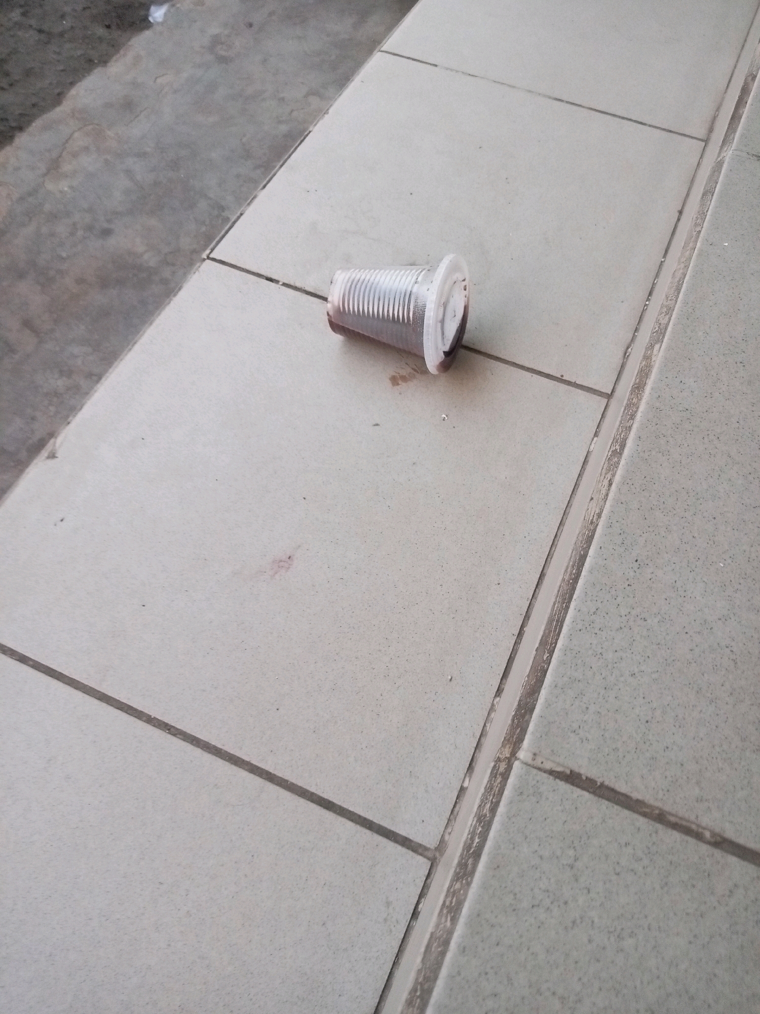 A clear plastic cup, with a white lid, lies on its side on a tiled surface. The cup appears to contain a dark liquid, possibly coffee or soda, and has some residue spilled around its base. The tiled surface is light-colored with darker grout lines, suggesting it is part of an outdoor patio or walkway. To the left of the tiled area, there is a rough, gray concrete surface, possibly a sidewalk or road. The overall scene suggests a common urban or public area in Batam, Indonesia, where discarded items might be found. There are no people or animals visible in the image.