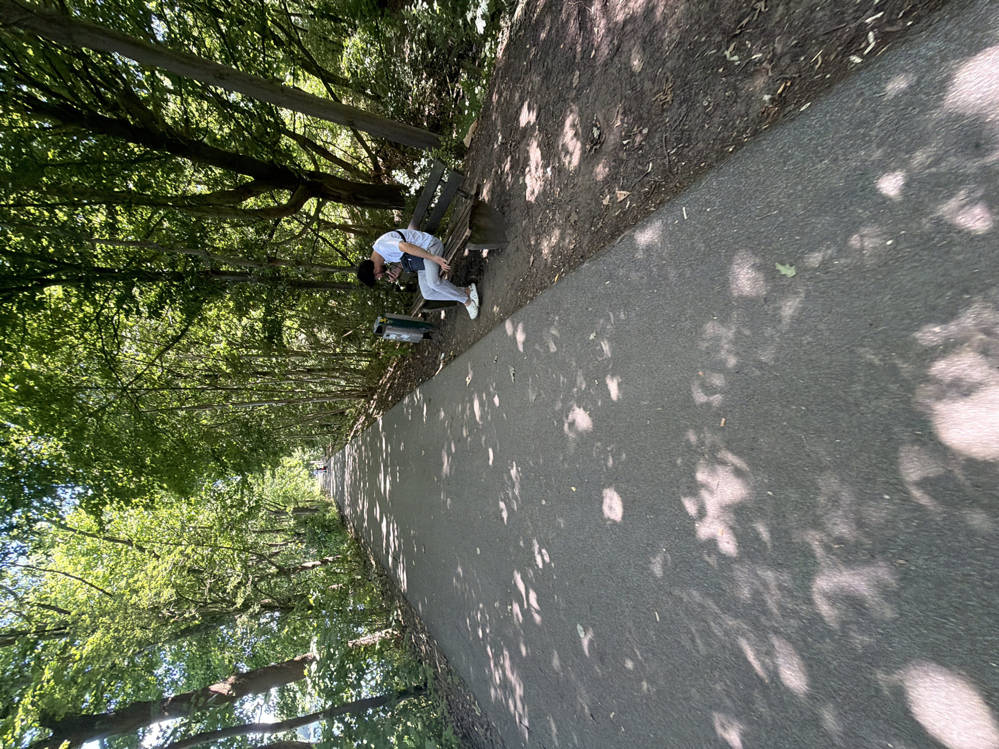 A person is sitting on a wooden park bench next to a paved path. They are wearing a white t-shirt, light grey pants, and white sneakers. A dark grey bag is slung across their chest. The person is leaning forward, looking down and touching their face, possibly with their hand. Next to the bench is a green trash bin.
The scene is outdoors, in a park or wooded area, with trees lining the path. The sunlight filters through the leaves, creating dappled shadows on the path and the ground. The trees are lush and green, suggesting it is daytime during a warm season. The path is grey asphalt. The ground next to the path is dirt and covered with leaves and debris.
The overall impression is one of a serene, natural setting. The time of day appears to be late morning or early afternoon, given the angle of the sun and the shadows. There is no visible text in the image.