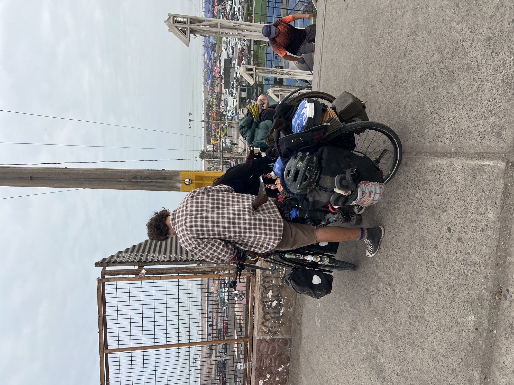 A cyclist is pictured on a bridge or elevated walkway in Arica, Chile. The cyclist is an individual with curly hair, wearing a checkered brown and white shirt and brown shorts. They appear to be adjusting items on their heavily-laden bicycle, which is packed with various bags and gear. The cyclist is standing next to a fence.
In the background, there is a dense urban environment with many cars and tents. At the higher level of the bridge, there is another person. The sky is overcast.