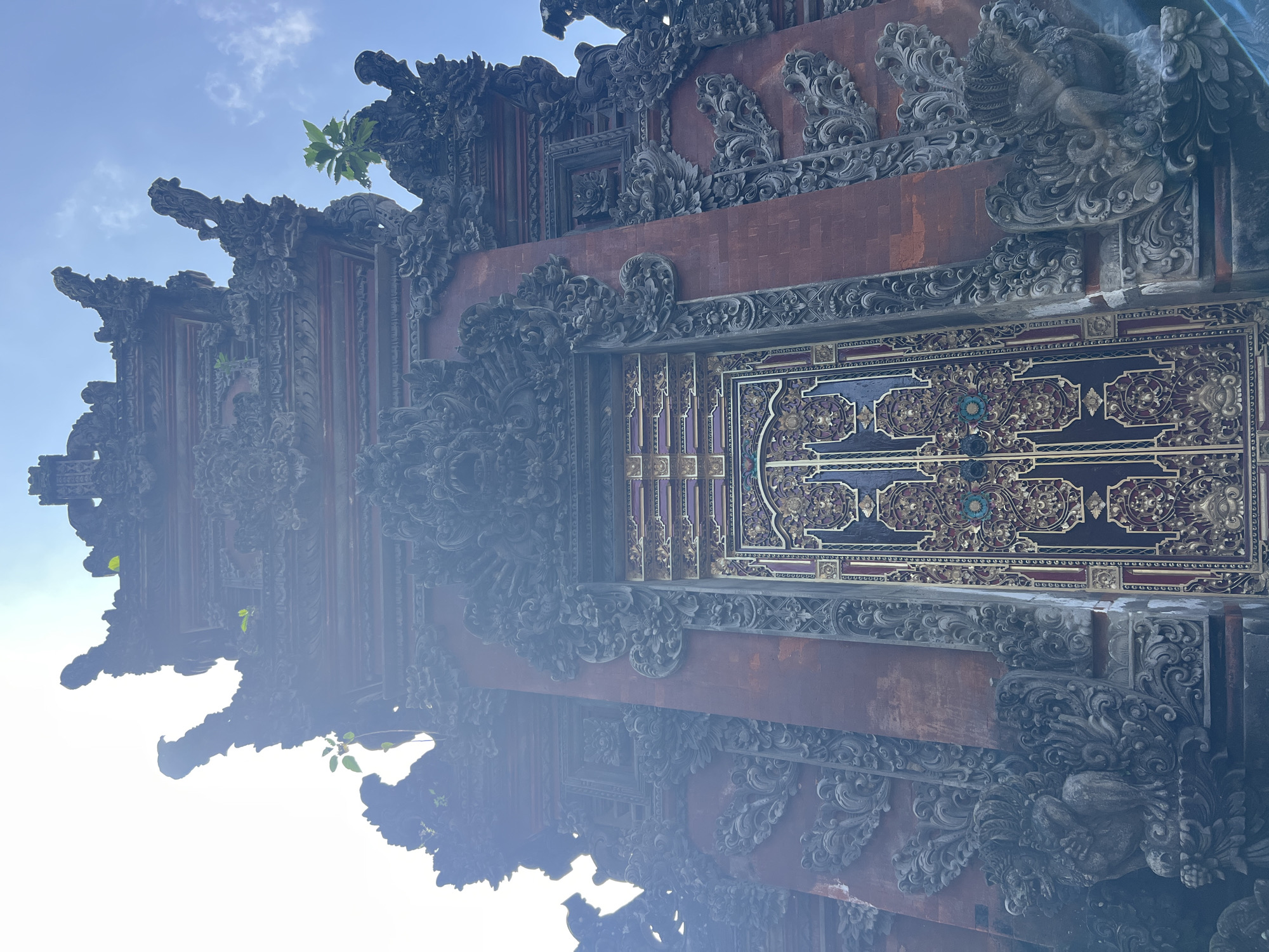 The image depicts a portion of a Balinese-style gate or doorway, characterized by intricate carvings and ornate decorations. The architectural structure is predominantly made of brick and dark gray stone or concrete, and adorned with elaborate floral and mythological motifs. A section of the door itself is visible, featuring a dark surface with gilded or painted embellishments in a floral pattern. Patches of brick are interspersed among the more detailed sections of the doorway. The photograph appears to have been taken looking upwards, capturing the sky partially visible in the upper and lower part of the frame. The style of the architecture and the presence of vegetation growing from the structure suggest this could be located in Sukawati, Indonesia, although this cannot be definitively confirmed without further information.