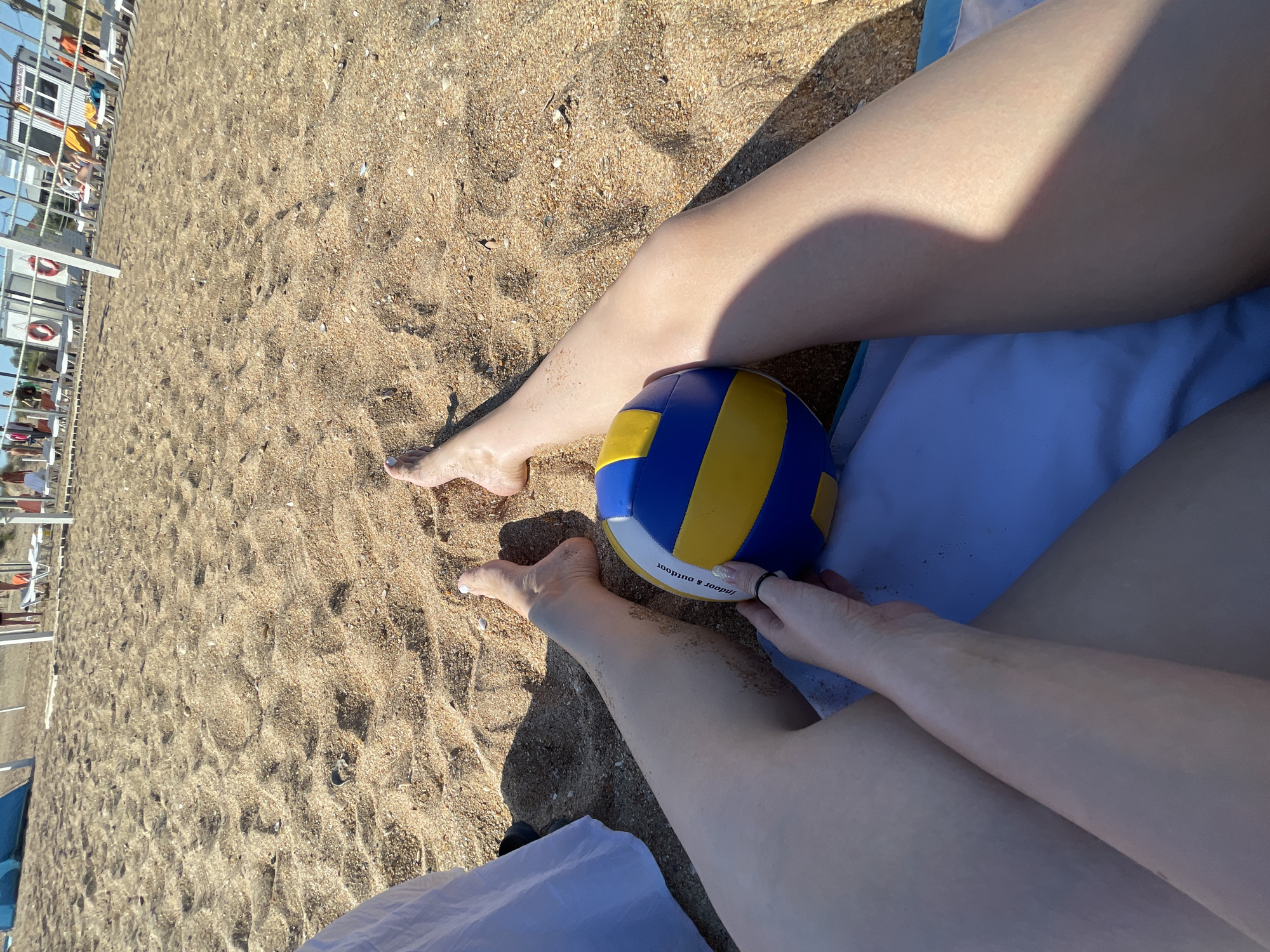 In Blagoveshchenskaya, Russia, a first-person perspective shot captures a segment of a person's legs and a volleyball on a sandy beach. The sand is a light brown color with visible shell fragments and footprints. The person's legs, fair-skinned and with some sand clinging to them, are positioned on a blue and white striped towel. A volleyball, predominantly blue and yellow with white sections, rests between their legs. The person's hand, with painted nails, is visible near the volleyball. In the background, other beachgoers and beach infrastructure, including lounge chairs and a building, are present, slightly out of focus. The scene is illuminated by bright sunlight, casting shadows on the sand.