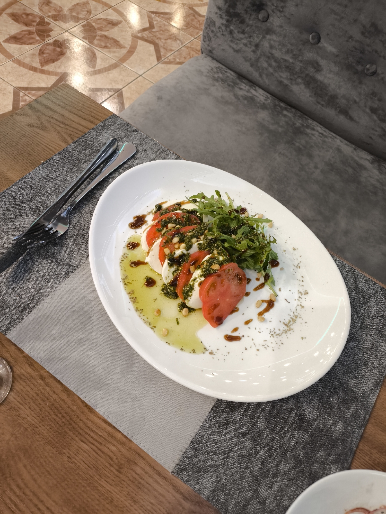 The image presents an inviting overhead view of a Caprese-style salad served on a white oval plate in a restaurant or cafe setting, located in Khimki, Russia.
The main subject is the meticulously plated salad, which consists of alternating slices of fresh mozzarella cheese and vibrant red tomatoes. It is generously garnished with a heap of fresh green arugula, drizzled with bright green pesto, and sprinkled with pine nuts. A clear, golden olive oil forms a pool at the base of the plate, enhanced with dried herbs, while dark, glossy streaks of balsamic glaze add both flavor and visual appeal.
The plate rests on a rectangular grey fabric placemat, which is situated on a warm-toned wooden table. To the left of the dish, a fork and knife are neatly placed, suggesting the meal is prepared for consumption. A small portion of another white dish is visible in the bottom right corner of the frame.
The background reveals a plush, dark grey upholstered booth or chair featuring button tufting, indicative of a comfortable dining environment. Beyond the table and seating, the floor is adorned with light-colored tiles, featuring an elaborate pattern of floral or star-shaped designs in a contrasting brownish tone. The lighting is bright and even, characteristic of an indoor restaurant setting. There are no people visible, and no text is discernible within the image. The overall scene evokes a serene dining experience, likely at mealtime.