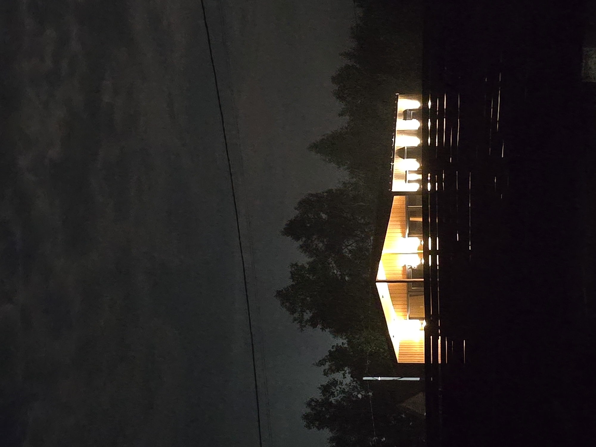 The image depicts a building illuminated by warm interior lights at night. The building has a gabled roof and vertical wooden siding. Multiple windows or openings are visible, each glowing with light, suggesting an indoor space. In front of the building is a dark, wooden fence or railing. Behind the building, silhouetted trees form a dark backdrop against a cloudy night sky. A thin wire stretches horizontally across the upper part of the image. The overall scene is dark, with the building being the primary source of light and visual focus. The location is identified as Ермолаево, Russia.