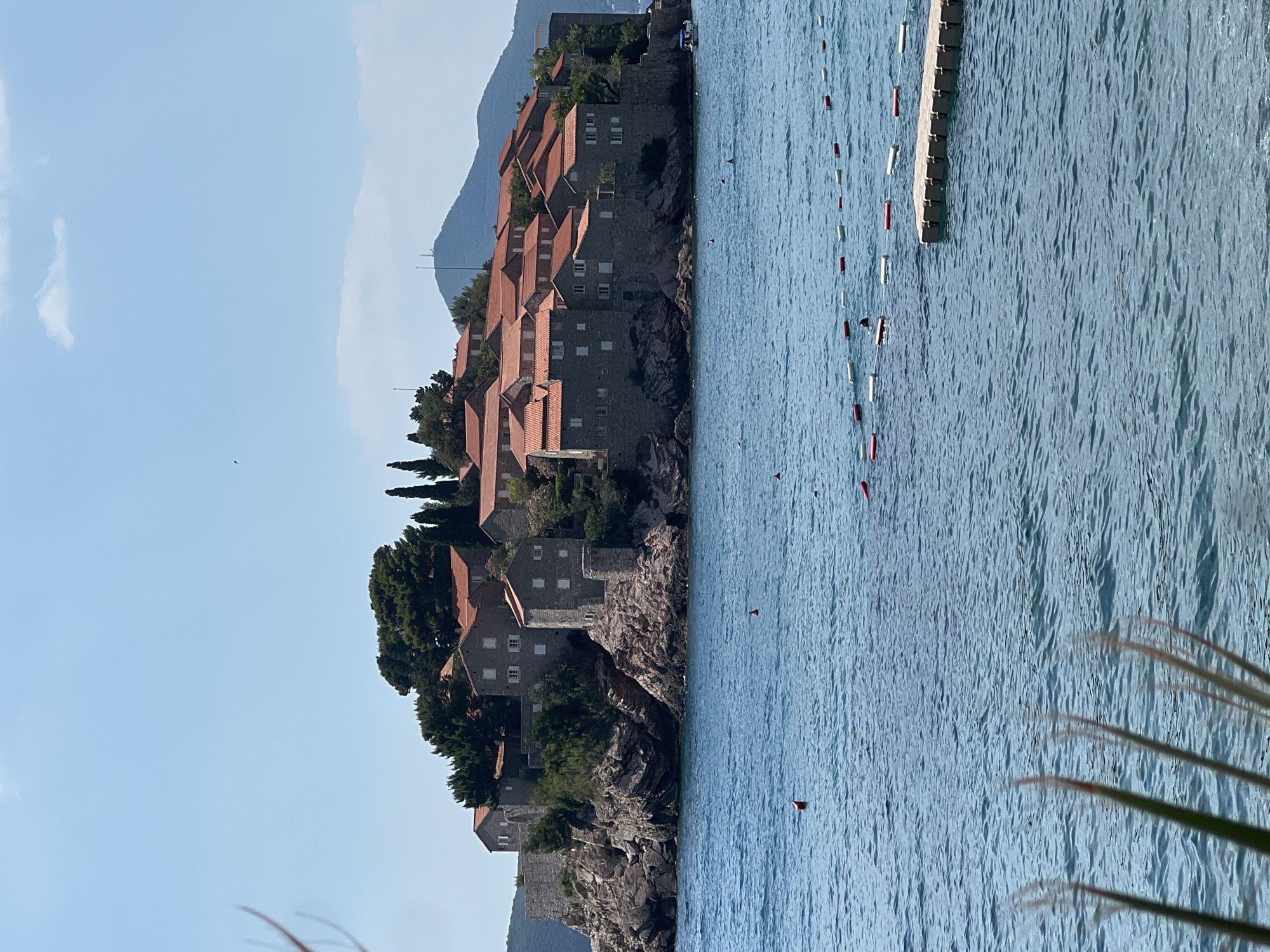 The image depicts Sveti Stefan, a small islet and fortified village on the Adriatic coast of Montenegro. The islet is connected to the mainland by a narrow causeway. The scene is dominated by the traditional stone houses with terracotta roofs that make up the village, perched on a rocky outcrop. Lush green trees are interspersed among the buildings, adding to the natural beauty of the setting.
The calm, bright blue water of the Adriatic Sea surrounds the islet. A series of buoys, some red and white, are visible in the water, marking a swimming area or perhaps a pathway for boats. A wooden walkway or pontoon extends into the sea from the right side of the image. The sky is a clear, pale blue, with a few wisps of clouds. The sun appears to be high in the sky, suggesting it is midday or early afternoon. The overall atmosphere is serene and picturesque, typical of a Mediterranean coastal landscape on a sunny day. There are no people visible in the image, and no text is present. The location is clearly identifiable as Sveti Stefan due to its distinctive island village architecture and natural setting.