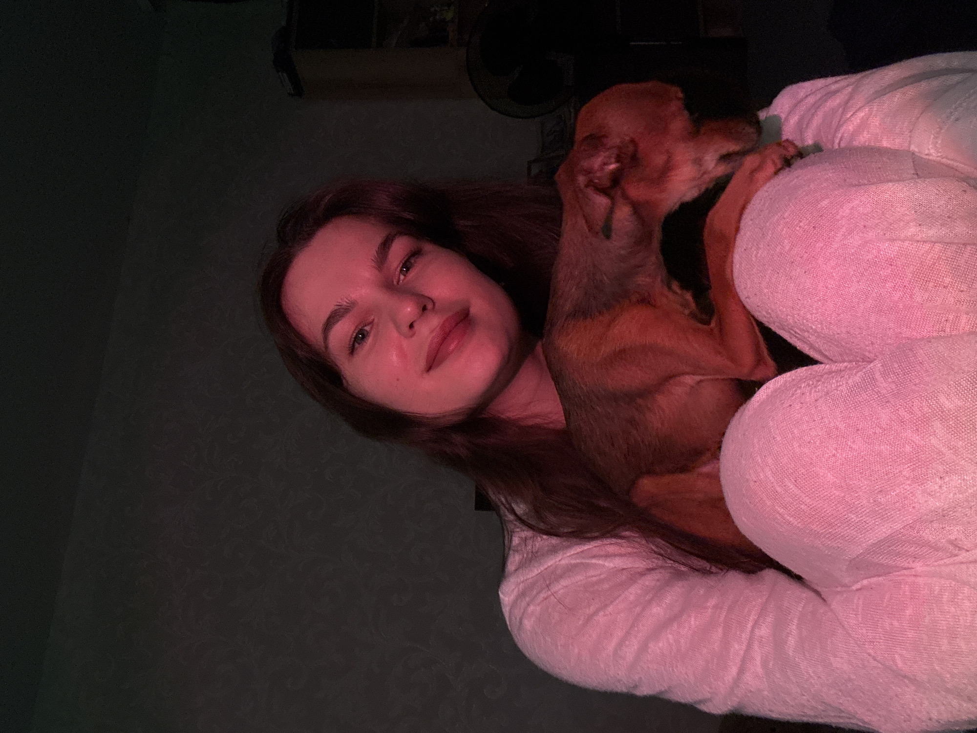 A young woman with long, dark brown hair and fair skin is captured in a close-up, indoor, selfie-style photograph. She is wearing a light pink, textured top. Resting on her lap and shoulders is a small, tan-colored dog. The lighting in the scene is dim and cast with pink and red hues, creating a soft, intimate atmosphere. The background appears to be a wall with a textured pattern. The woman is looking directly at the camera with a gentle smile, while the dog is looking down and away. The image is devoid of any specific landmarks or elements that would indicate a location within the city of Chaikovsky, Russia.