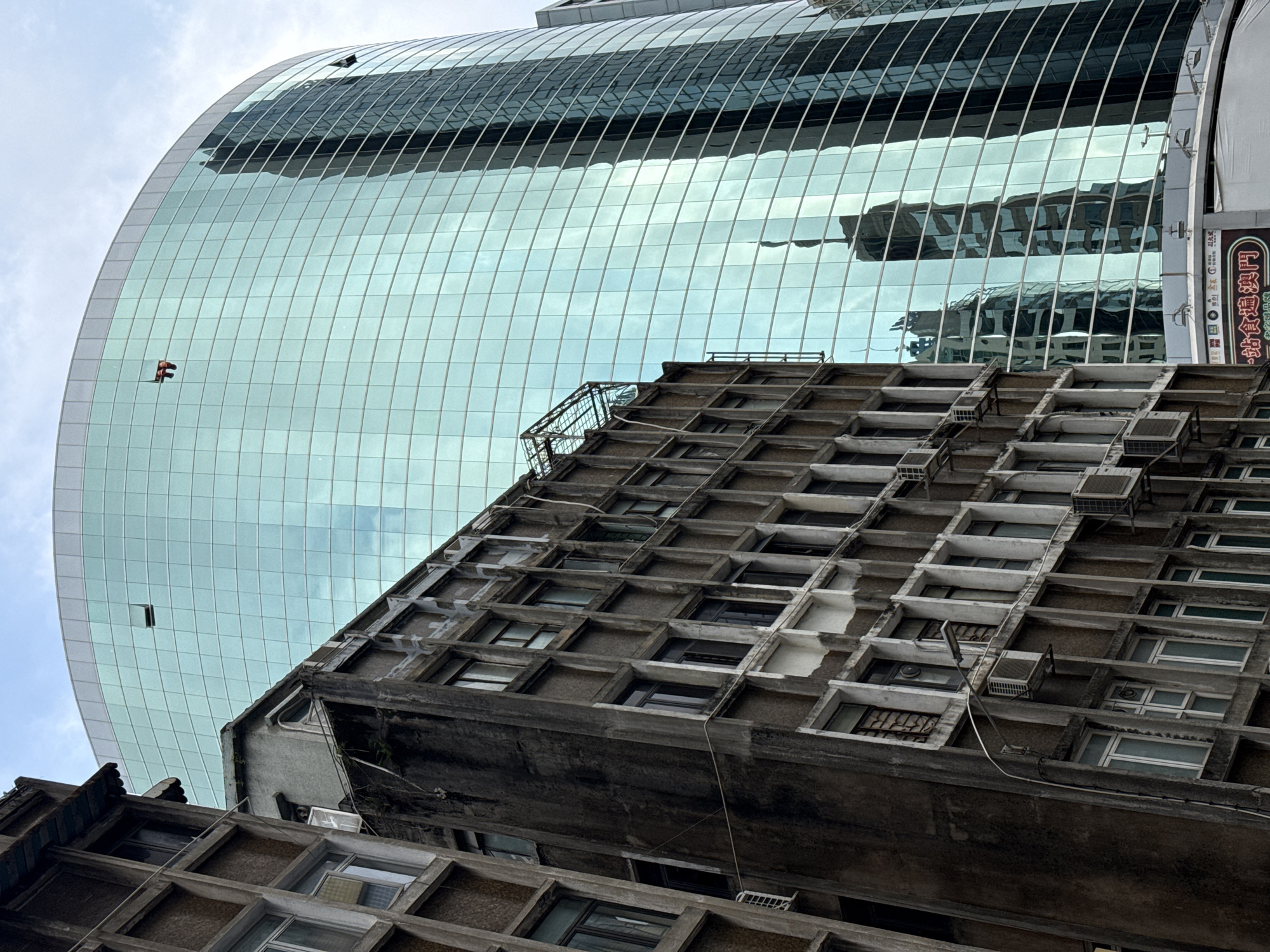 The image depicts a juxtaposition of architectural styles, with a modern, rounded glass building reflecting the environment above a more traditional, weathered concrete structure. The concrete building features a repeating pattern of recessed windows, many of which have individual air conditioning units installed. The angle suggests a low vantage point looking upwards. The reflective surface of the glass building mirrors surrounding structures, including another building with similar window patterns. A sign with Chinese characters is visible on the right side of the frame. This image was taken in the city of Macau, China.
