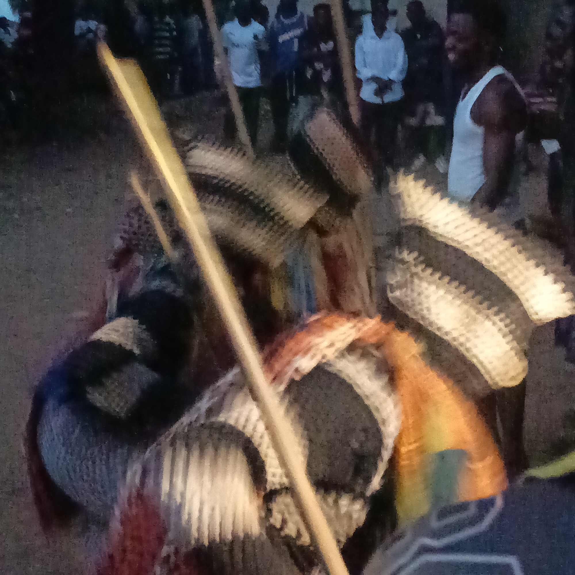 Akwanga, Nigeria. The image captures a chaotic scene featuring masked individuals, possibly participants in a performance or ceremony, with striped masks and attire, some holding staffs. Background includes a group of onlookers. One individual wears a white tank top, while others are clad in t-shirts. The environment appears to be an outdoor gathering at dusk.