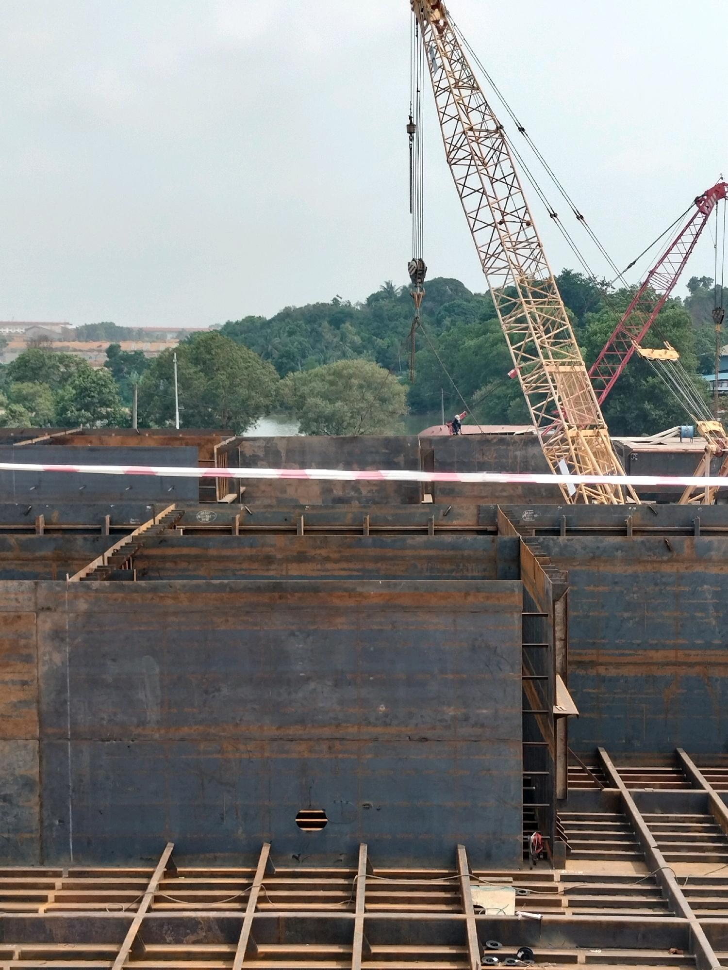 The image depicts a construction site in Batam, Indonesia, likely at a shipyard or industrial facility. Dominating the scene are large, dark steel structures, possibly components of a ship or a large industrial building, arranged in a grid-like formation. These are supported by a framework of wooden beams. Two large cranes are prominently visible. A yellow lattice crane is positioned in the mid-ground to the right, its boom extending upwards. To its right, a red lattice crane's boom is also visible. A single person, wearing dark clothing and a hard hat, is standing on the steel structure near the yellow crane. The environment appears to be outdoors, with lush green trees and a body of water visible in the background, suggesting a waterside location. The sky is overcast and light.