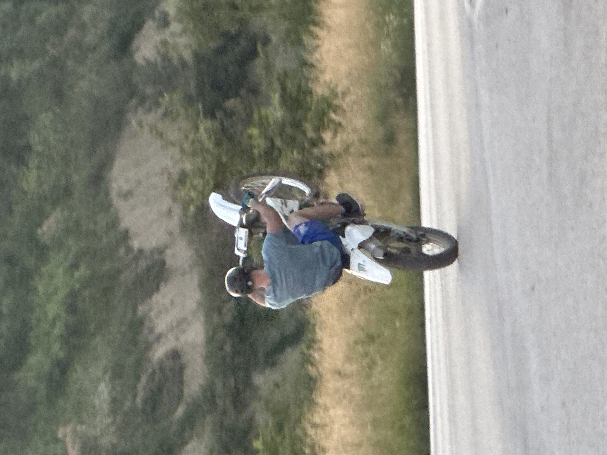 A person in a blue t-shirt and blue shorts is performing a wheelie on a white dirt bike on a paved road. The person is wearing a dark helmet and black shoes. The dirt bike's front wheel is elevated off the ground. The background consists of a blurred green and brown hillside with sparse vegetation. The road surface appears to be asphalt. The image quality is low, with visible graininess and a lack of sharp detail. The specific location in Prisovo, Bulgaria cannot be determined from the visual information provided.