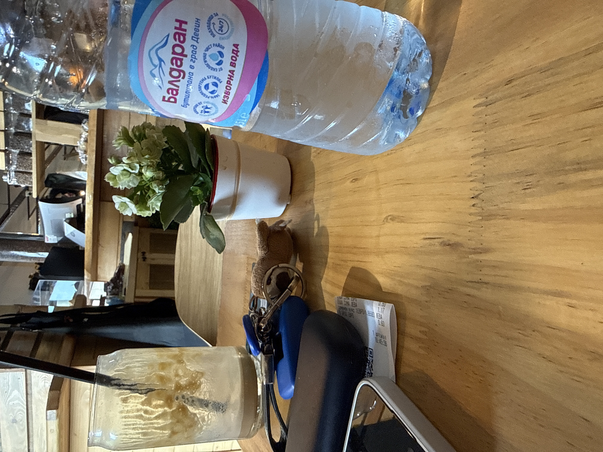 The image, oriented in portrait (rotated 90 degrees counter-clockwise), presents a tabletop scene from what appears to be a cafe or restaurant in Kardzhali, Bulgaria. The surface is a light-colored wooden table.
A clear plastic bottle of "Балдаран" (Baldaran) spring water is a central object. Its label, in Bulgarian, identifies it as "бутилирана в град Девин" (bottled in the town of Devin) and "ИЗВОРНА ВОДА" (Spring Water). Other text indicates it's a "100% рециклируема бутилка" (100% recyclable bottle) and "подходяща за децата" (suitable for children). Beside the bottle, a small white potted plant with delicate light green flowers provides a decorative element.
Further down, a tall glass holds an iced, light brown beverage, likely coffee or tea, with a dark straw. Next to the glass are a set of keys, featuring a small brown teddy bear keychain and two blue keyfobs. A dark-colored mobile phone or its case is partially visible nearby.
A crumpled paper receipt lies on the table, displaying Bulgarian text such as "ОБЩА СУМА" (Total Amount) and the currency "ЛЕВА" (Leva), along with prices and a timestamp of "16:45:36", indicating late afternoon. The background is softly blurred, showing vague wooden structures and interior details, suggesting a public indoor setting. No individuals are visible, but the arrangement implies a person's recent presence or imminent return.