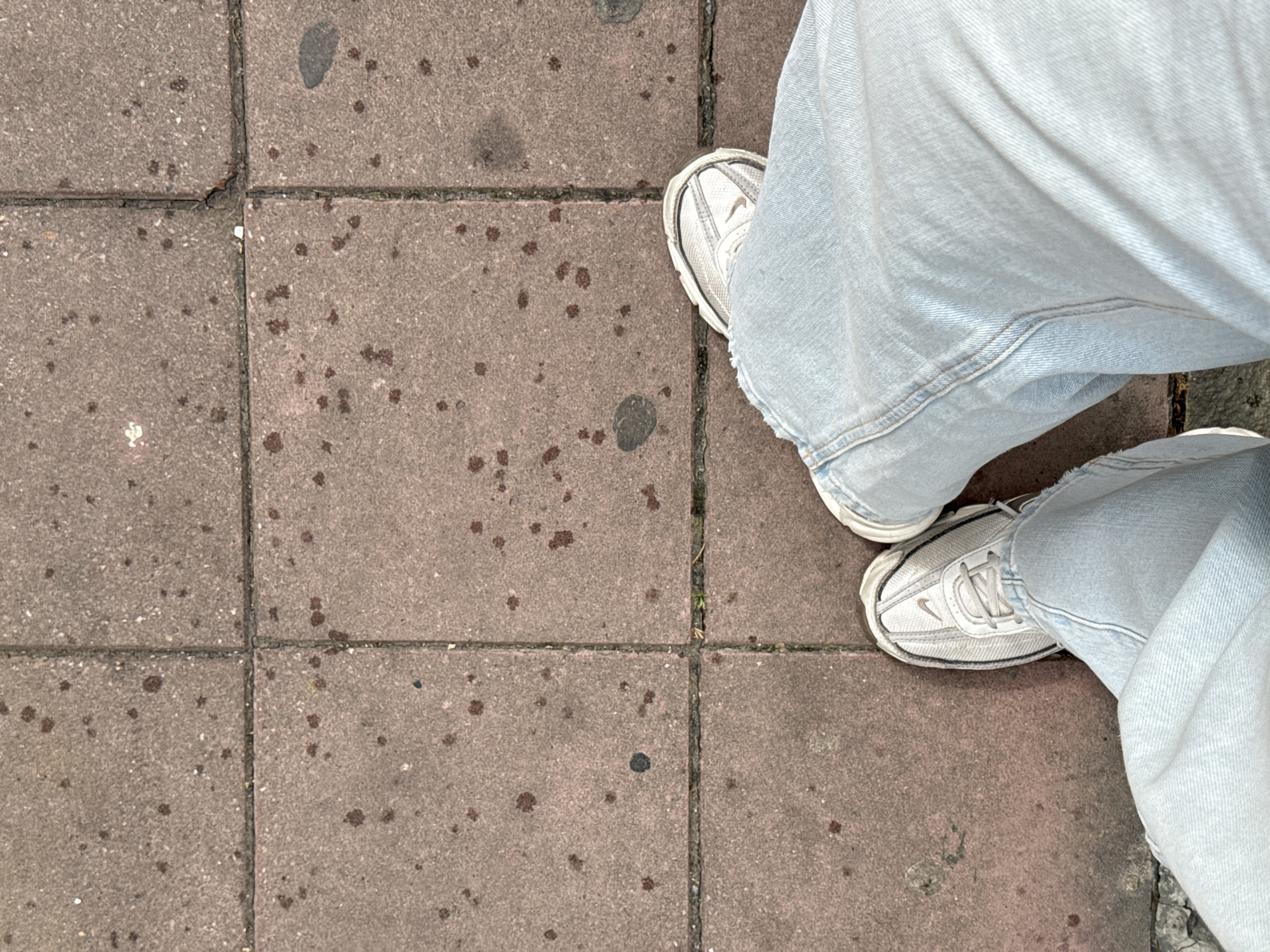 A first-person perspective image depicts a person standing on a sidewalk made of square, reddish-brown concrete tiles. The tiles are marked with dark spots of an indeterminate substance. The person is wearing light blue denim jeans and white sneakers with a visible Nike swoosh logo. The location is indeterminate.