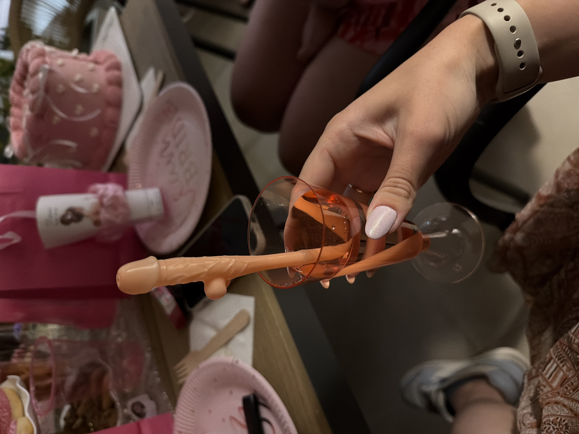 A hand with light pink nail polish holds a champagne flute filled with a light pink beverage. A portion of a phallic-shaped novelty object is also visible, resting on the rim of the flute and extending outward. The scene appears to be a party, suggested by the presence of a pink cake with white icing decorations, pink paper plates with "BRIDE" printed on them, and small packaged items. The setting is indoors or outdoors with a casual, celebratory atmosphere, possibly a bridal shower or bachelorette party. There are other people in the background, though they are out of focus. The overall mood is festive and playful.