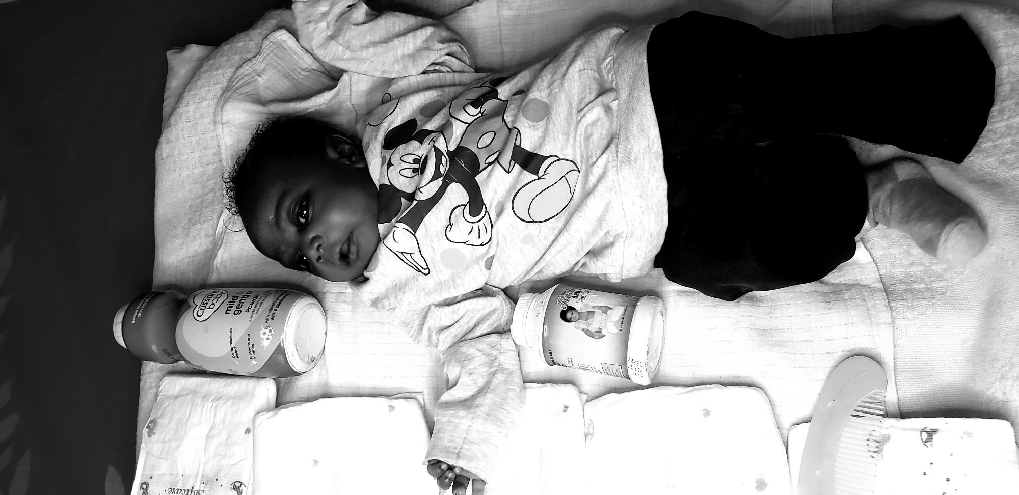 The image depicts an infant lying in a crib. The baby wears a light-colored top with a Mickey Mouse design and dark pants. The crib contains diapers, baby powder, and a comb. The location is Narayi, Nigeria, based on the baby powder brand.