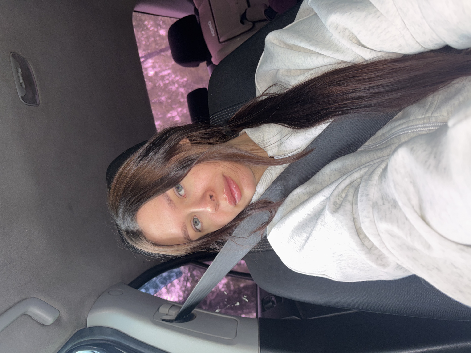 A young woman is in the front passenger seat of a car. She is wearing a light gray hoodie and has dark brown hair with a blonde streak. She is wearing a seatbelt. The background shows the interior of the car and a window with trees visible outside. Based on visual cues alone, it's impossible to determine if the image was taken in the city of Chaikovsky, Russia. The interior of the car is gray and black. There is a child seat in the back. The woman is looking at the camera.