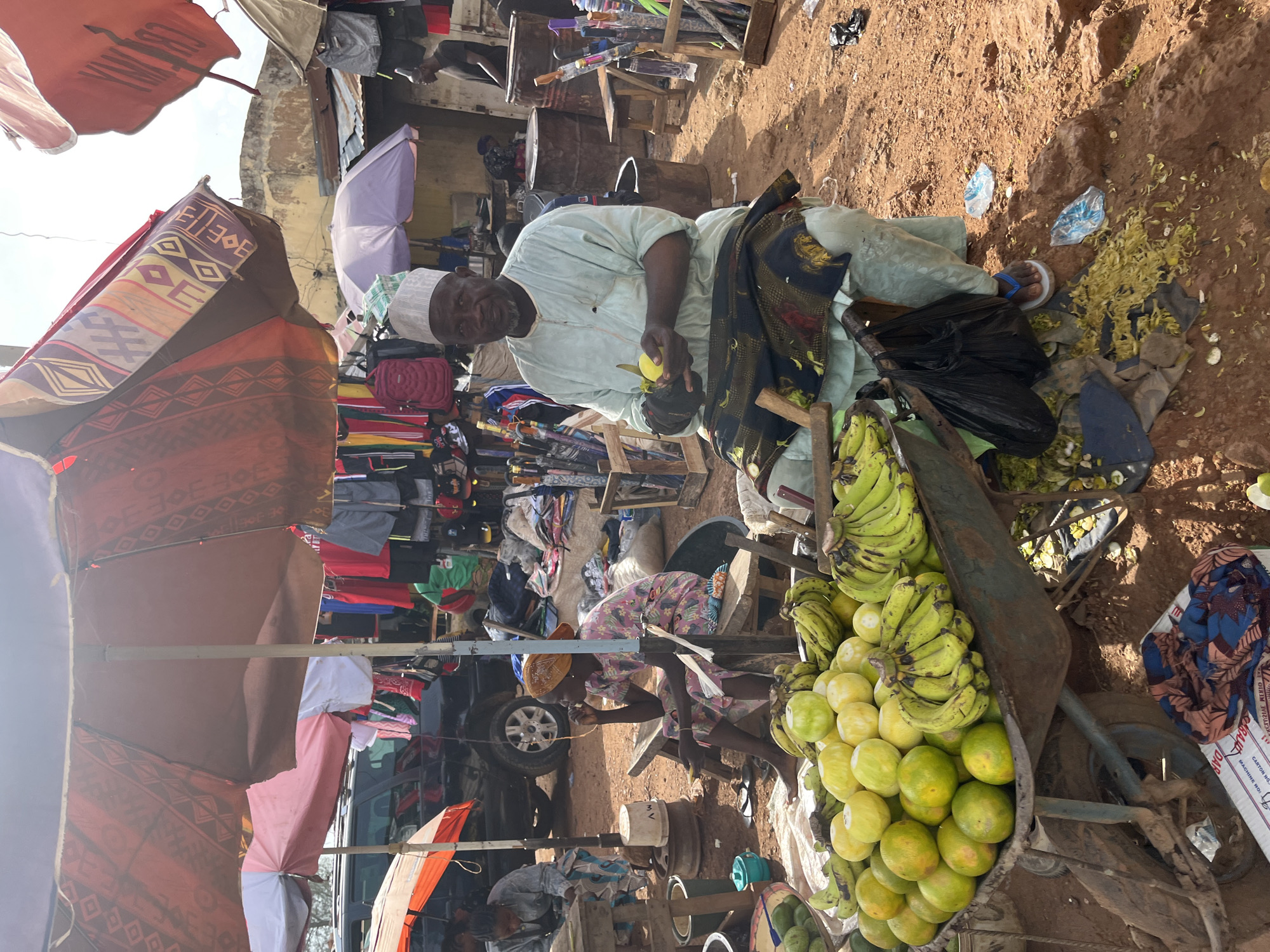 This image depicts an outdoor market scene in Rafin Par, Nigeria. The main subjects are two individuals, likely vendors, amidst stalls displaying various goods. One man, wearing a light-colored robe and a white cap, is seated and holding a yellow fruit and a knife, suggesting he might be preparing produce. He is positioned near a cart overflowing with bananas and other citrus fruits. The other individual, a woman wearing a patterned dress and a headscarf, is also seated, seemingly engaged with something in her hands.
The setting is a bustling market with a variety of items for sale, including clothing, bags, and umbrellas, displayed under colorful awnings. The ground is covered in dry, reddish soil, indicative of a sunny environment. Scattered debris, including discarded fruit peels and plastic wrappers, suggests the ongoing activity of the market. The time of day appears to be late morning or early afternoon, given the bright sunlight casting shadows.
The overall atmosphere seems busy and functional, characteristic of a local marketplace. The vendors appear focused on their tasks, with no overt expressions of emotion visible. In the background, there are structures that suggest permanent stalls or buildings, interspersed with makeshift displays. The presence of a vehicle in the middle ground implies it might be part of the market infrastructure or a delivery vehicle. There is no visible text that can be clearly deciphered in the image.
