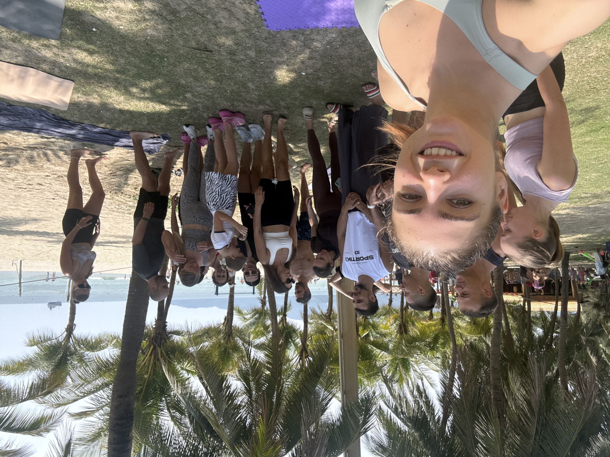 A group of approximately 15 people, mostly women, are posing for a selfie on a beach. The selfie is being taken by a woman in the foreground, who is smiling. The others are standing behind her, mostly facing the camera. They appear to be in athletic wear or casual clothing.
The beach is lined with palm trees, and in the background, there is an ocean with a small island or peninsula visible. There is also what appears to be a boat on the water.
Based on the vegetation, beach, and architectural elements visible, it is likely that the location is My Khe Beach, in the city of Đà Nẵng, Vietnam. In the background there is what appears to be a beachside structure or small building. Some yoga mats are partially visible on the ground.