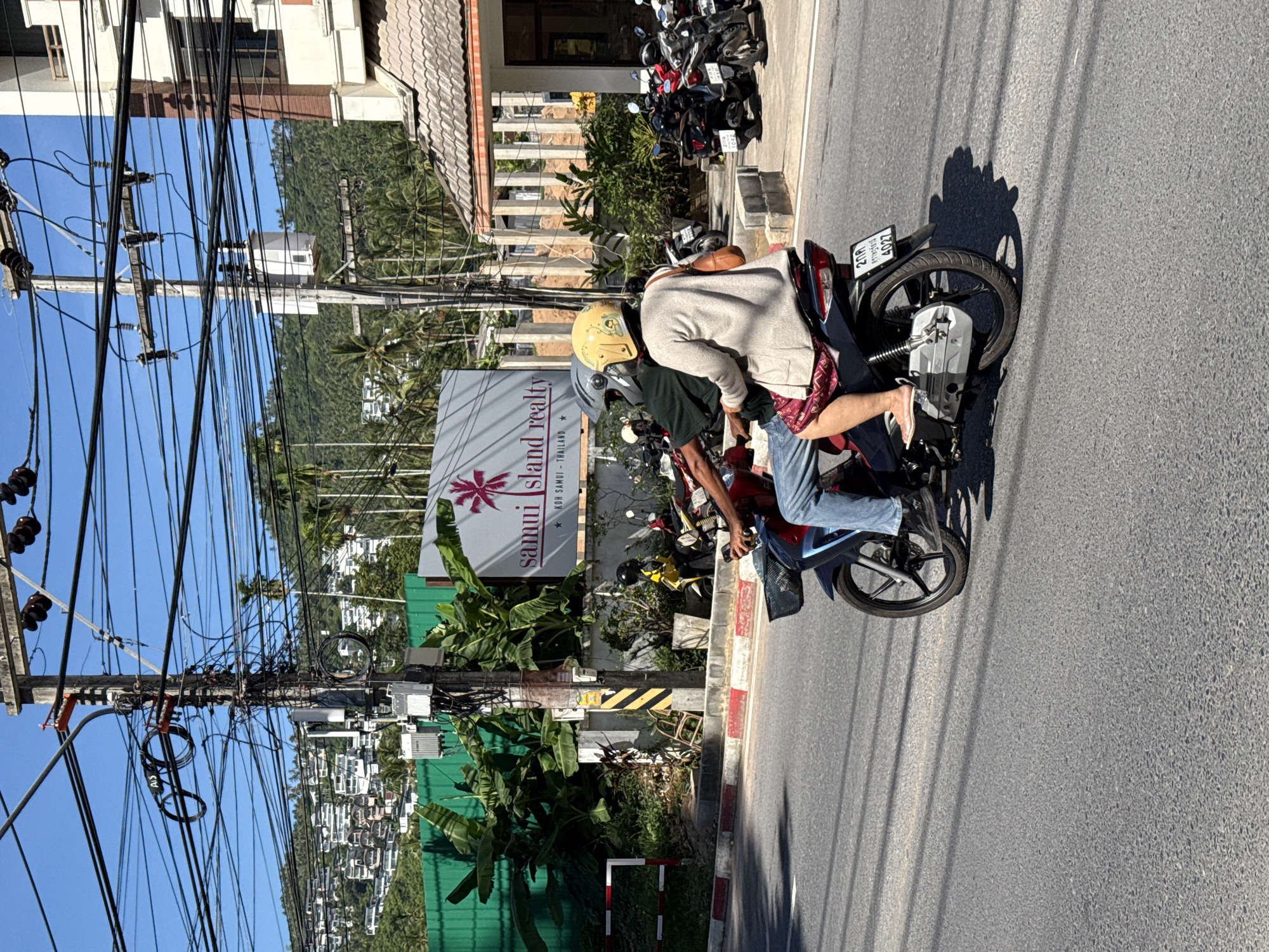 The image depicts a street scene in Baan Chaweng Noi, Thailand, under a clear blue sky. In the foreground, two individuals are riding a motorcycle on an asphalt road. The rider, wearing blue jeans, is partially visible, while the passenger, a woman with a yellow helmet and a light-colored cardigan, is seated behind. She is wearing a patterned red skirt and has a tan shoulder bag. The motorcycle has a black basket on the front and a license plate with Thai characters and the number "4027".
In the background, a sign for "samui island realty" is visible, with a palm tree logo. Adjacent to the sign is a green construction barrier. A utility pole, heavily laden with numerous electrical wires, stands prominently to the left of the sign, with various electrical components attached. Beyond the barrier and buildings, a hillside covered with dense tropical vegetation and numerous houses is visible. Several parked motorcycles are also present near a building on the right side of the image. The overall environment suggests a tropical urban or suburban setting with significant overhead utility infrastructure.